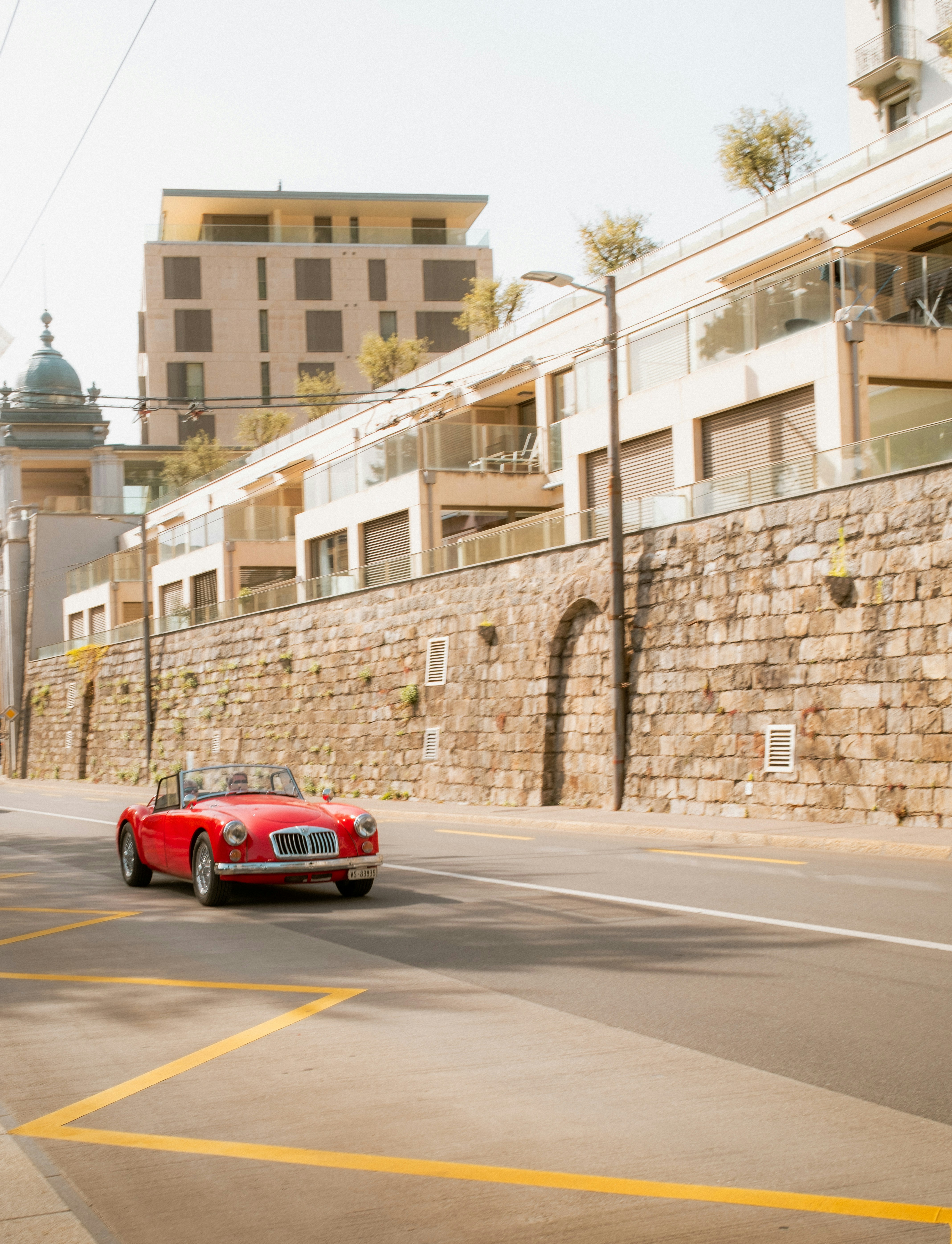 a-red-car-driving-down-a-street-next-to-a-stone-wall-jQ4GWcJ360U