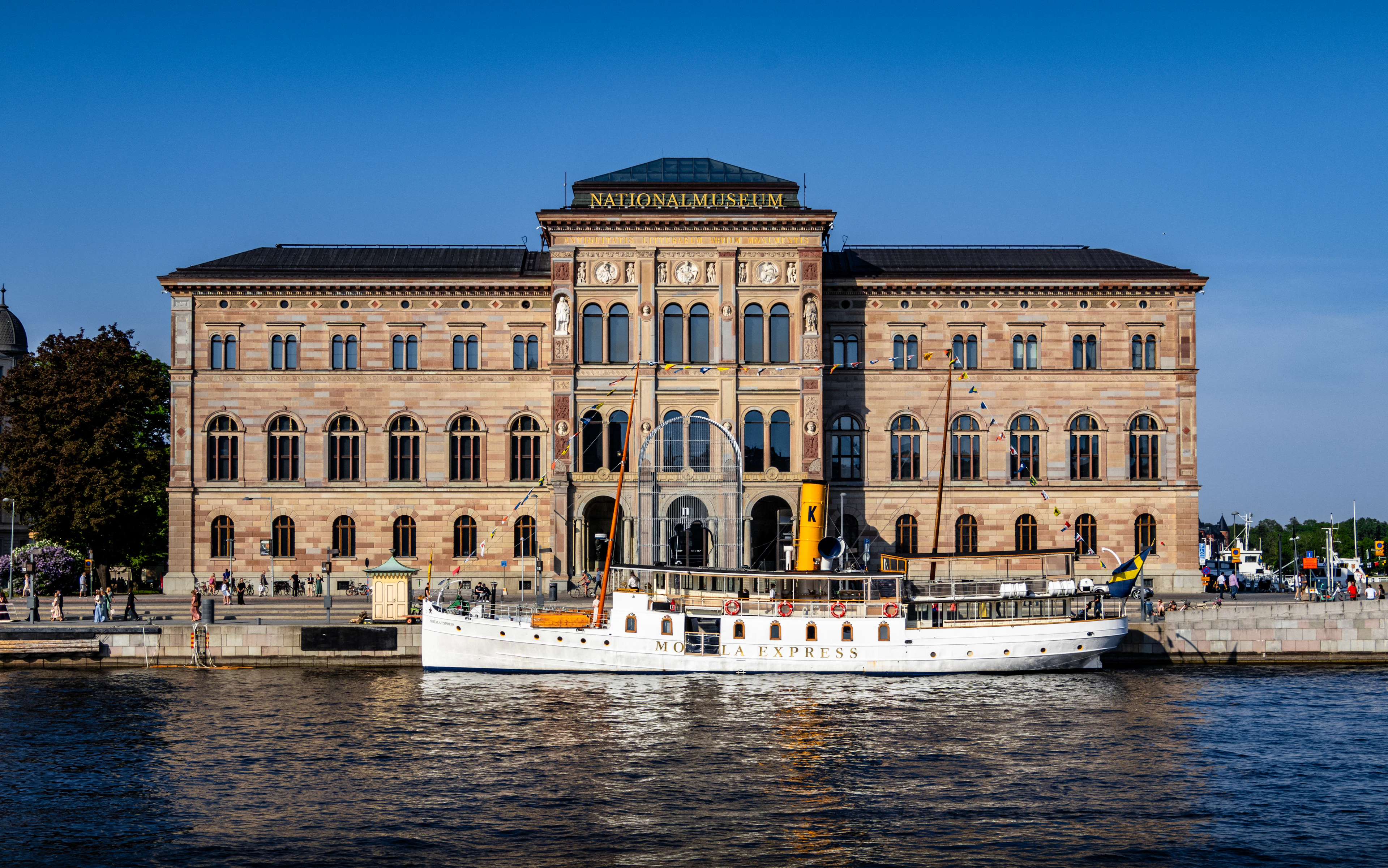 The sandstone Nationalmuseum on Stockholm's Blasieholmen waterfront in strong summer sun, a white heritage steamer moored along the quay in front of it under a deep blue sky.