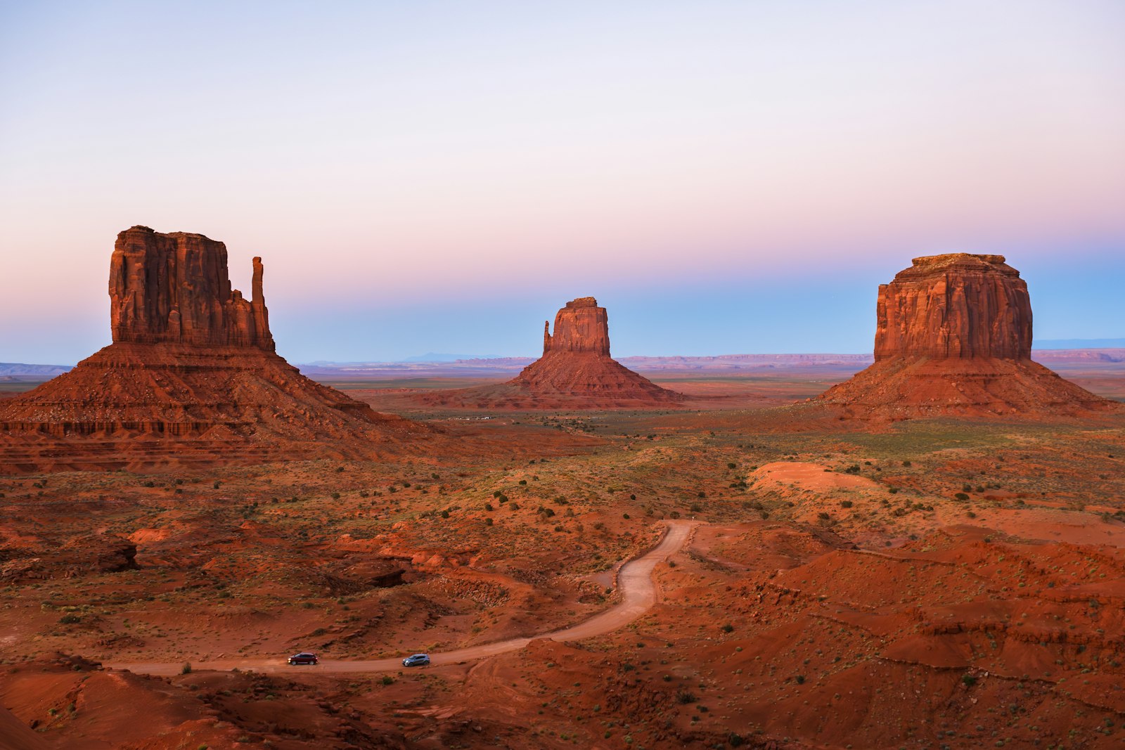 A landscape photograph showing three prominent red sandstone buttes (the Mittens and Merrick Butte) in a desert valley with a dirt road and two vehicles in the foreground.
