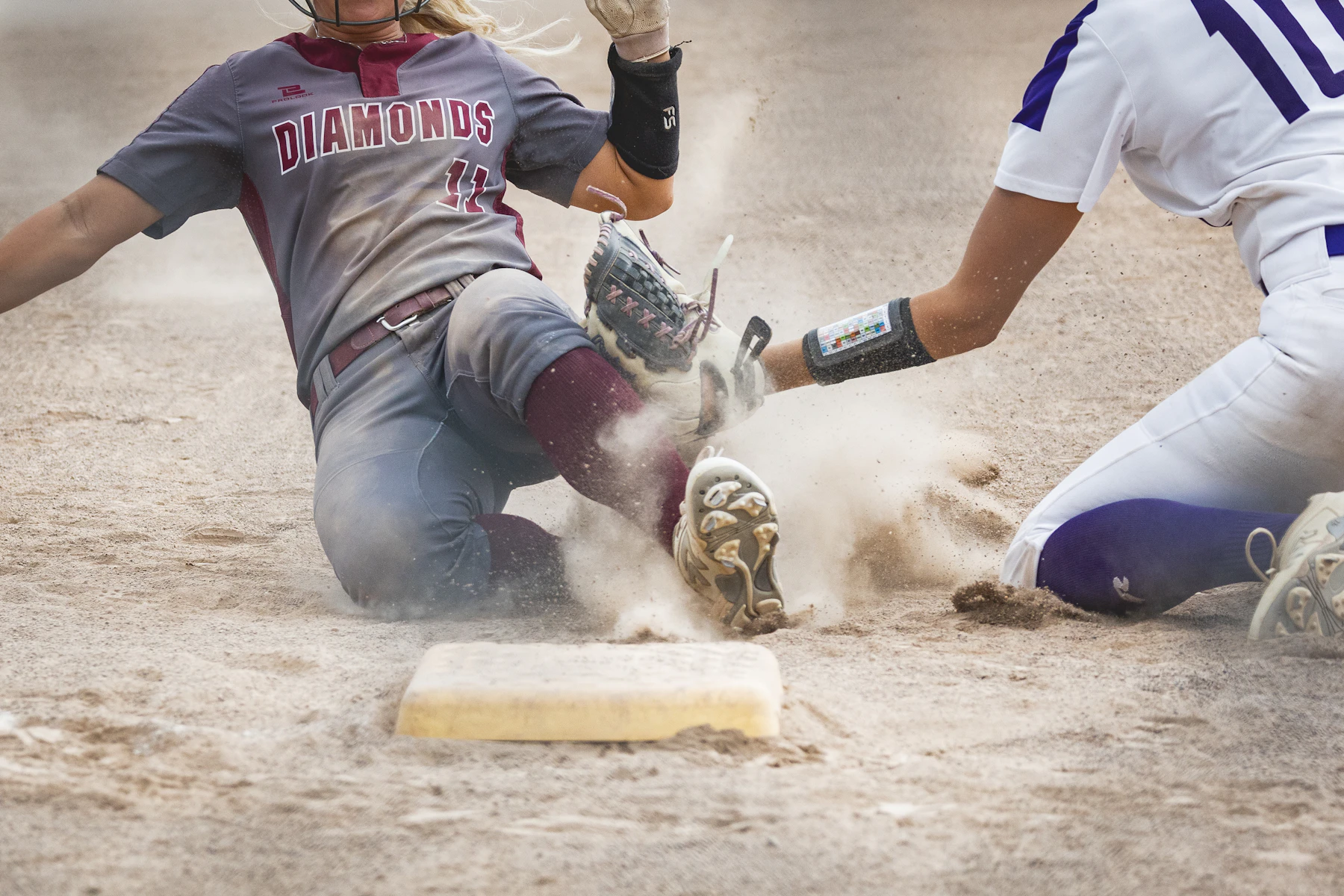 A softball runner sliding feet first into a base in a cloud of dirt as a fielder reaches to tag