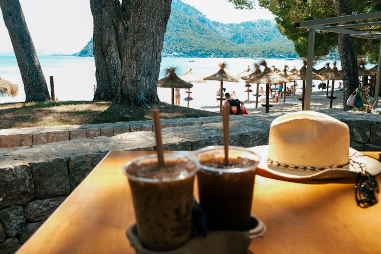 a table with two drinks and a hat on it looking toward the bay of pollensa mallorca