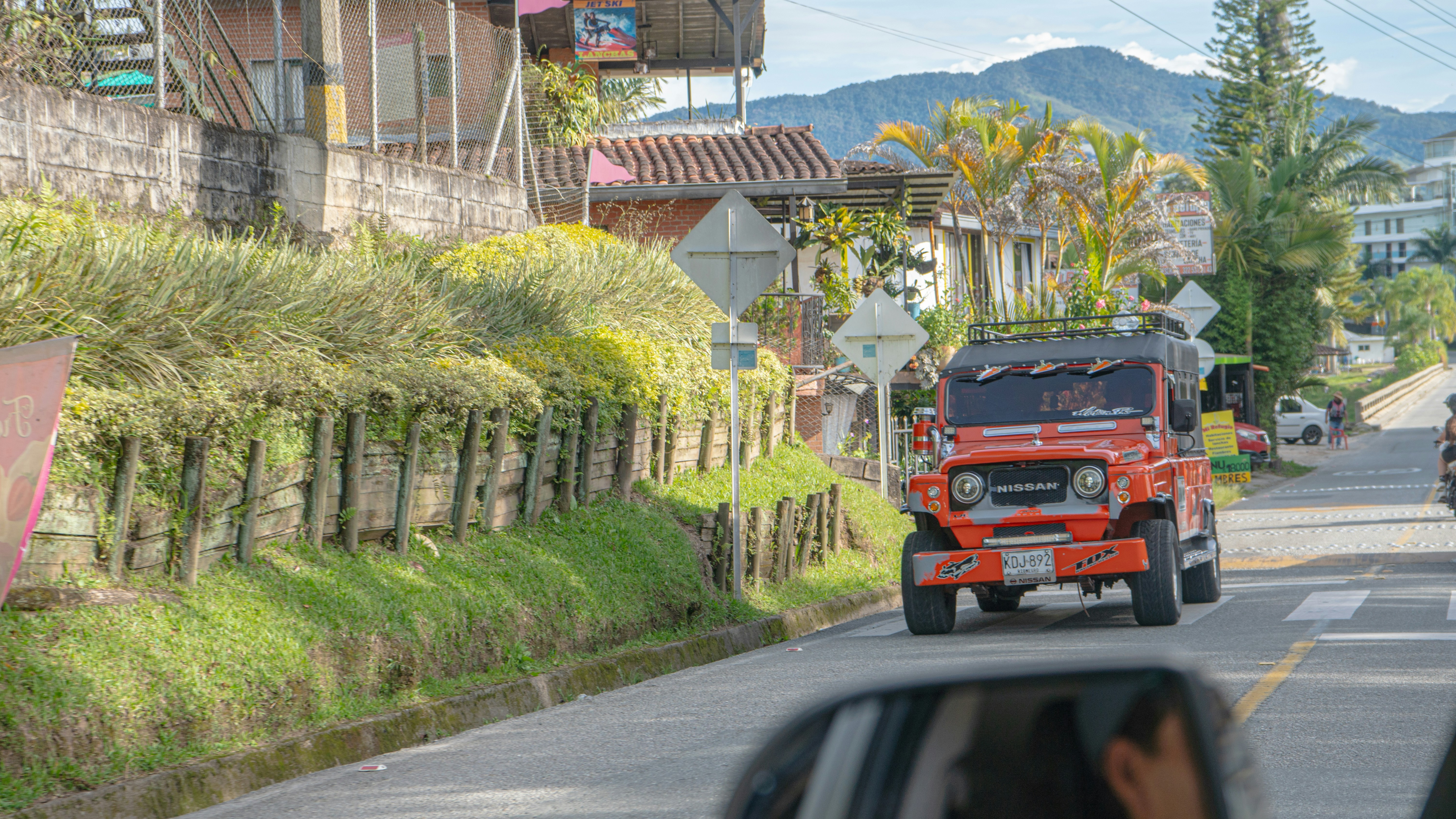a red jeep driving down a street next to a lush green hillside