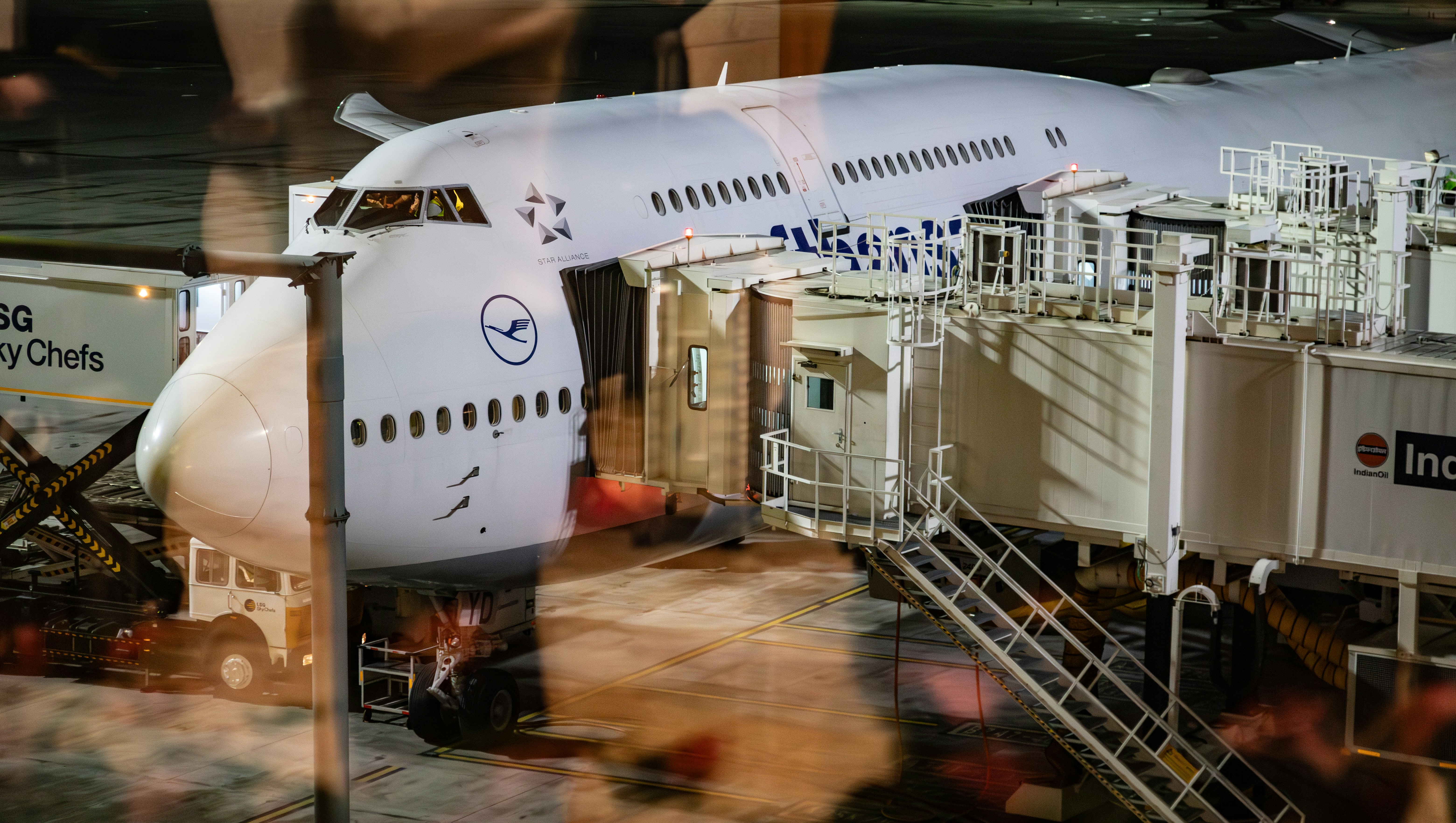 a large jetliner sitting on top of an airport tarmac, This image shows a close-up view of a large Lufthansa airplane docked at an airport terminal during nighttime. The aircraft is connected to a jet bridge, with LSG Sky Chefs