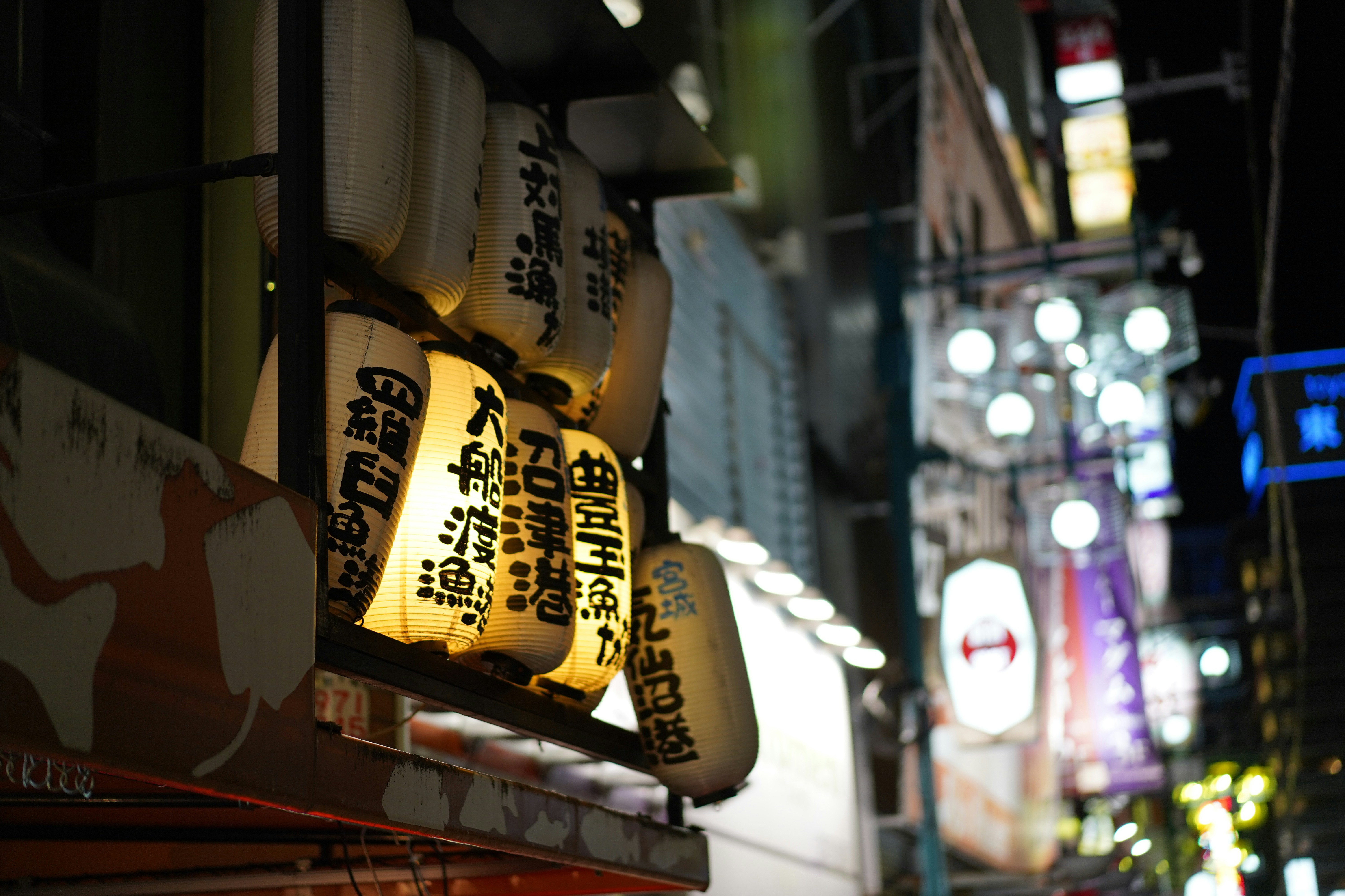 a bunch of lights that are on the side of a building, This captivating photograph taken in Osaka showcases a vibrant street scene illuminated by traditional Japanese lanterns. The warm glow of the lanterns contrasts beautifully with the surrounding neon signs and city lights, creating a dynamic and atmospheric night scene. The composition highlights the blend of modern urban life with traditional cultural elements, typical of Osaka