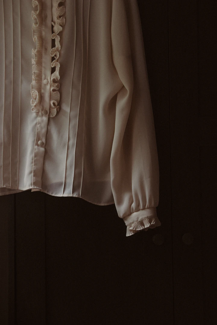 Still-life photograph of a bone-coloured ruffled blouse hanging on a hook