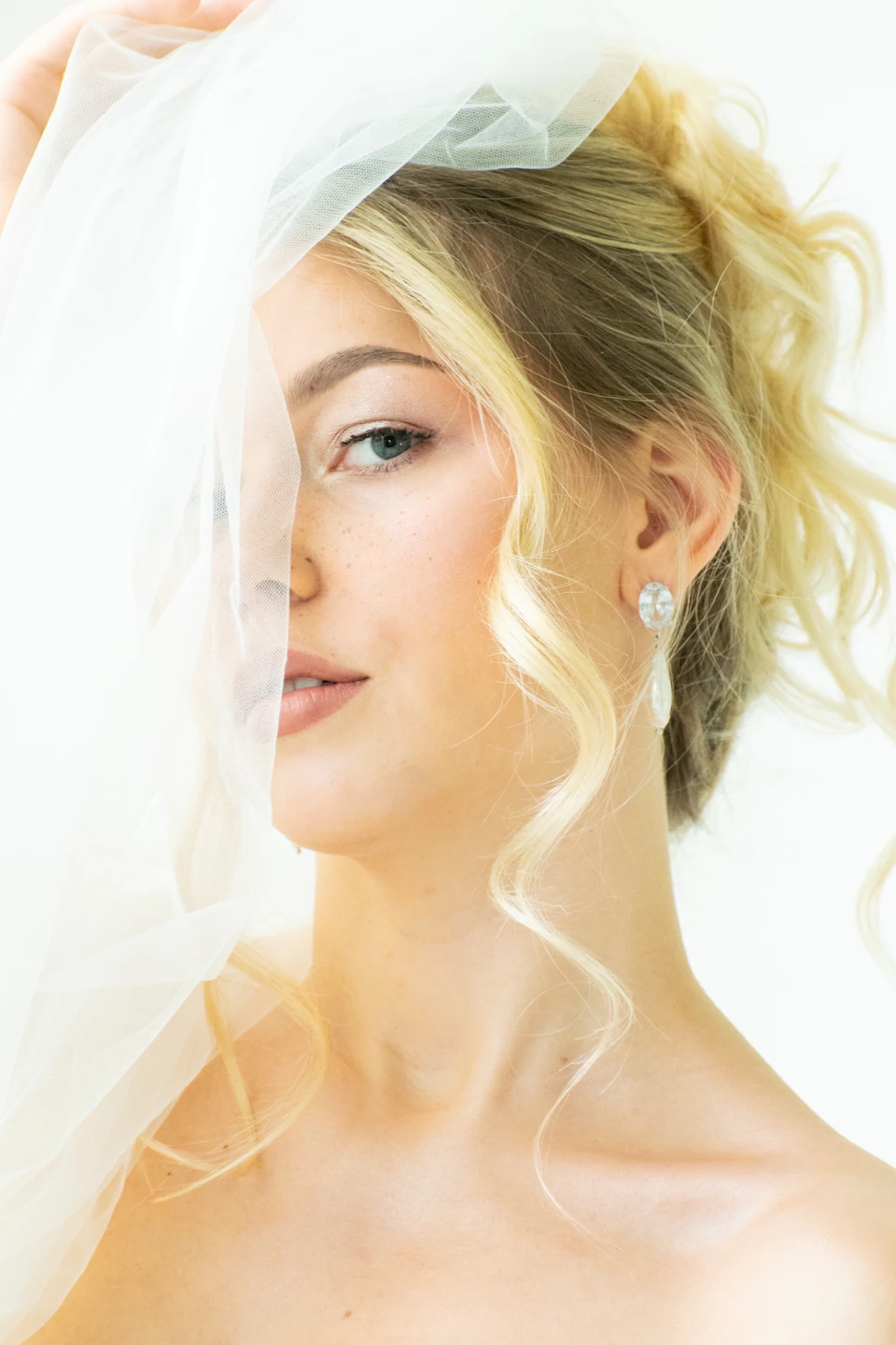 The image shows a close-up view of a woman's face, with her hair styled in loose curls and wearing a white veil that partially covers her eyes. The background is blurred, drawing focus to the woman and her attire.