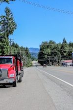 A red truck driving down a street next to a forest