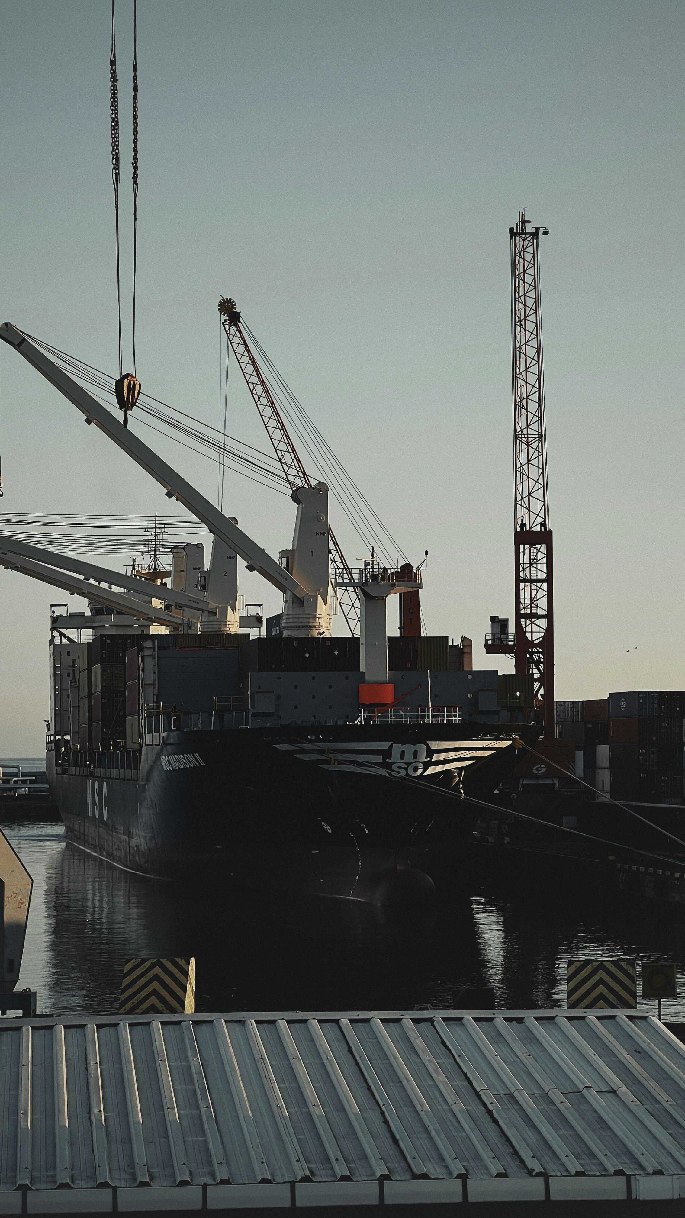 Container ship at port with cranes loading cargo, moody overcast sky, deep shadows, industrial port lighting at dusk