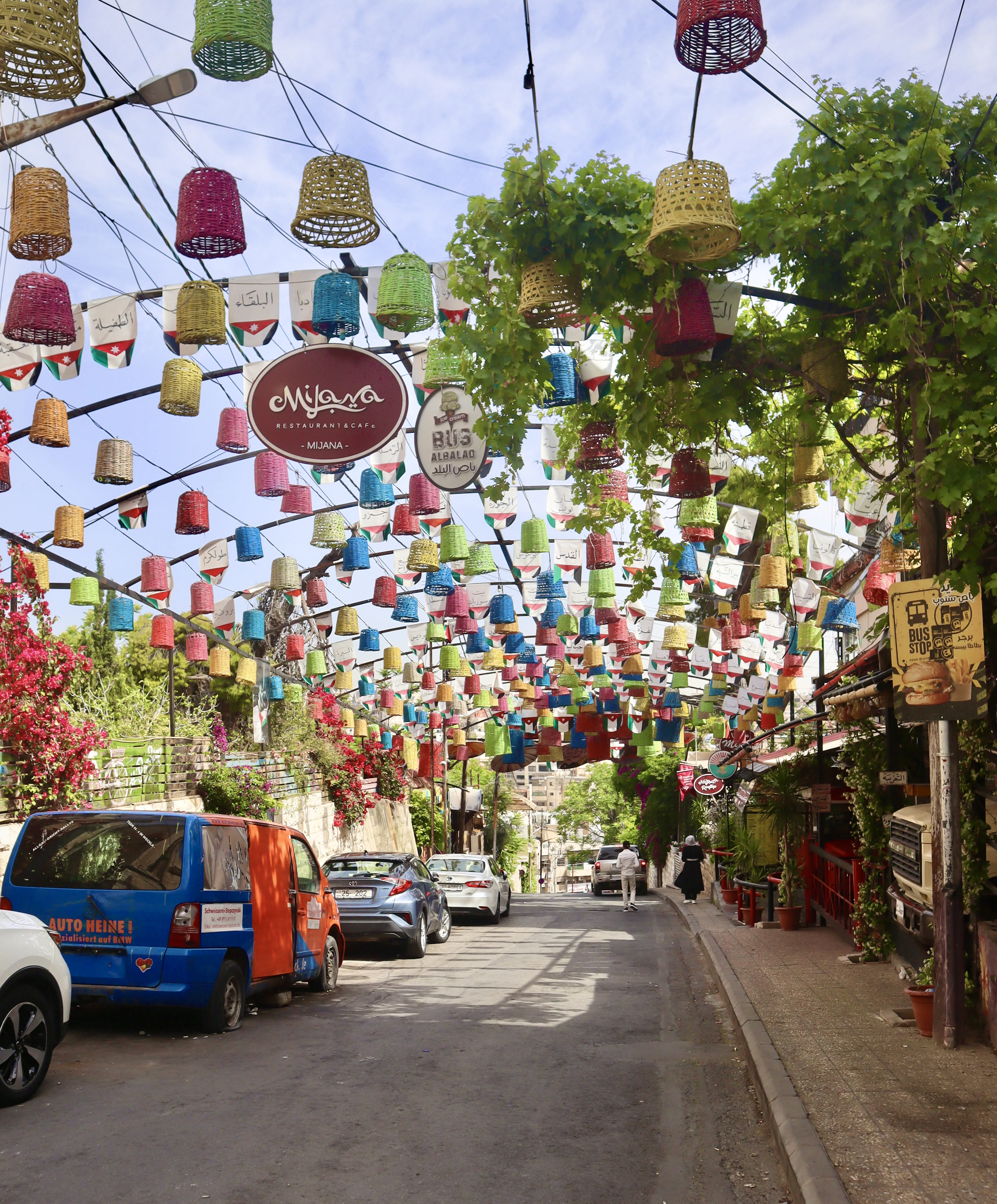 Colorful hanging lanterns and baskets canopy a lively, plant-lined street in Amman's Rainbow Street district.