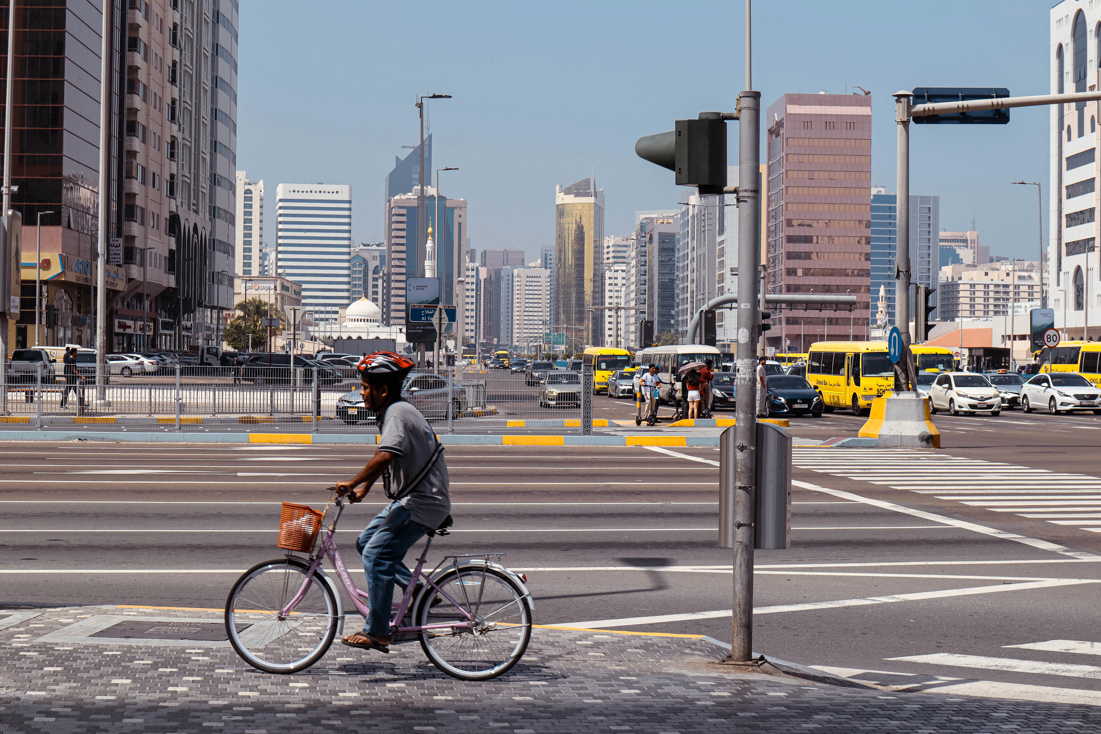 A man rides a bicycle across a wide Abu Dhabi junction in bright midday sun, yellow taxis and city buses lined up behind him and the downtown towers rising beyond.