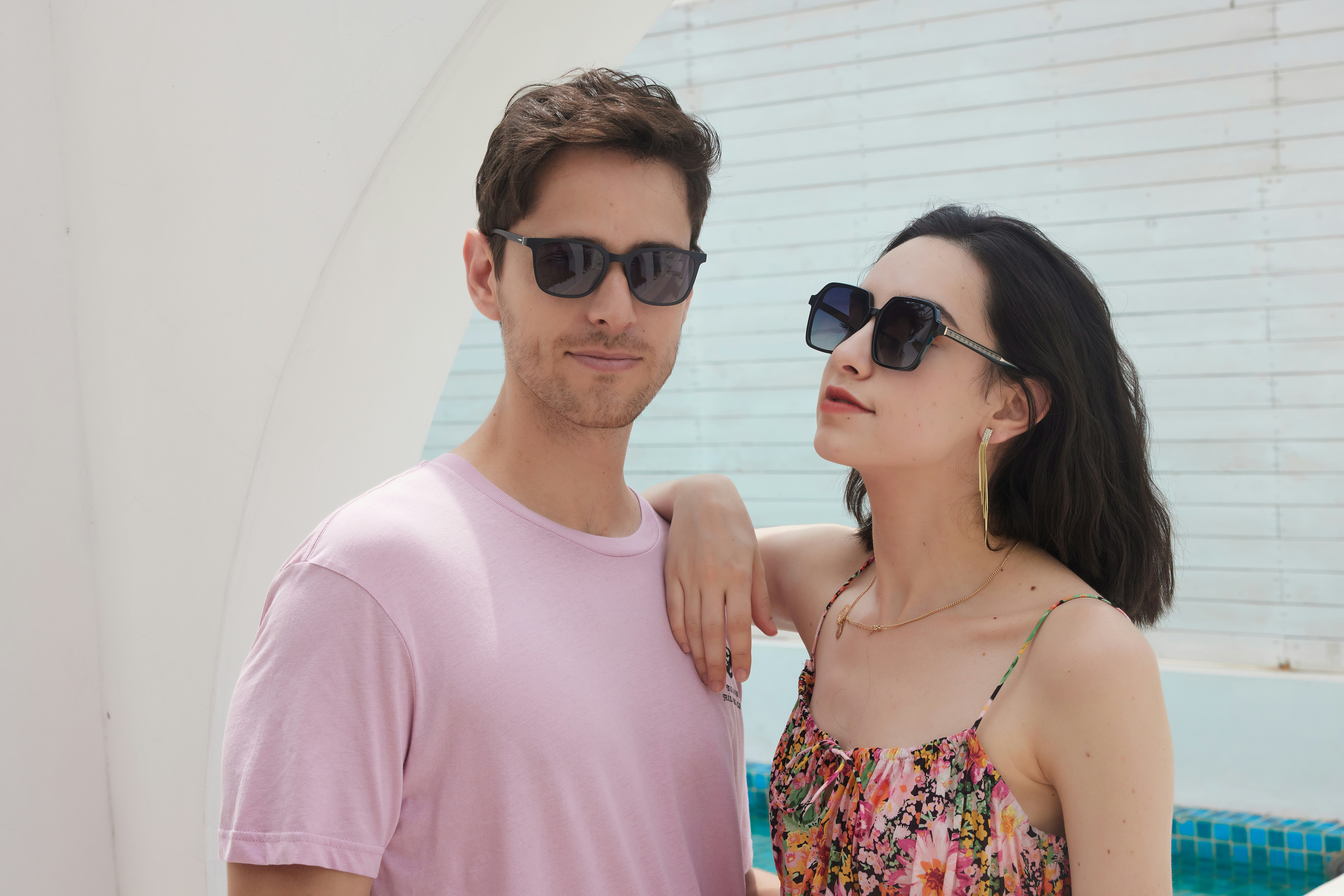 A man and a woman standing next to each other, A stylish couple exudes confidence and charm, showcasing their fashionable sunglasses. The man, in a light pink t-shirt, and the woman, in a colorful floral dress, pose effortlessly against a modern, minimalist backdrop. Their coordinated look and relaxed demeanor highlight the versatility and contemporary appeal of their eyewear.