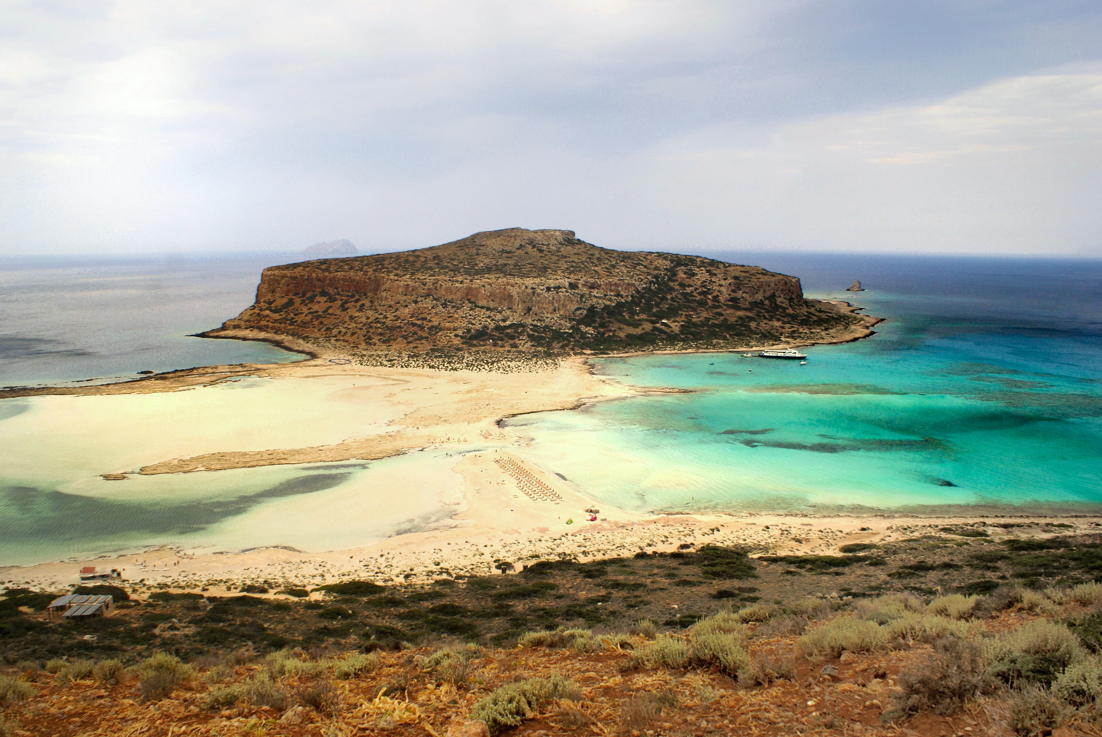 The empty sandbar and turquoise shallows of Balos lagoon on Crete's north-west tip, the flat rocky headland of Cape Tigani rising beyond, under a soft grey and gold overcast sky