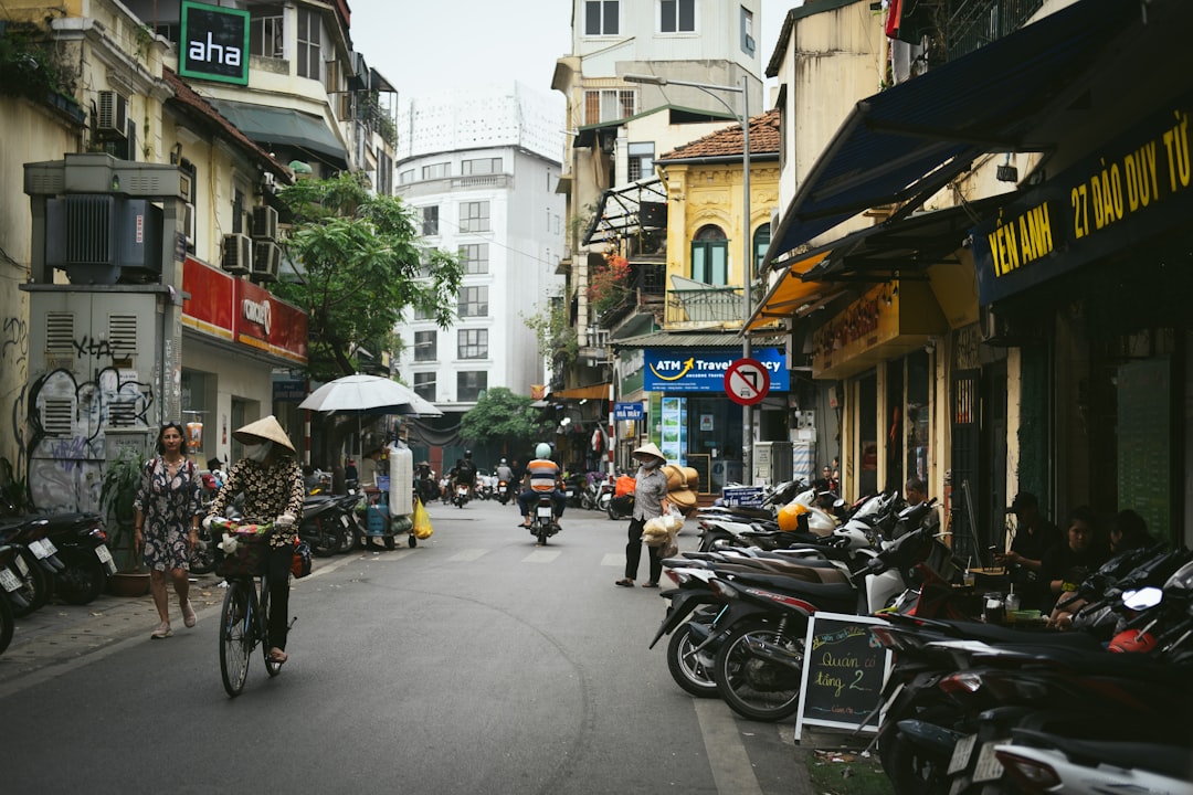 A group of people riding bikes down a street, A typical street in Hanoi, bustling with activity. The narrow street is lined with motorbikes parked on the sides, and a few cyclists and pedestrians navigate the road. Traditional buildings with shops and eateries on the ground floor add to the vibrant urban atmosphere. The architecture reflects a mix of old and modern influences, with signs in Vietnamese and lush greenery providing a backdrop. People are seen wearing casual clothing, and a woman in a conical hat rides a bicycle, highlighting the blend of tradition and modernity. The scene captures the daily life and dynamic culture of Hanoi.