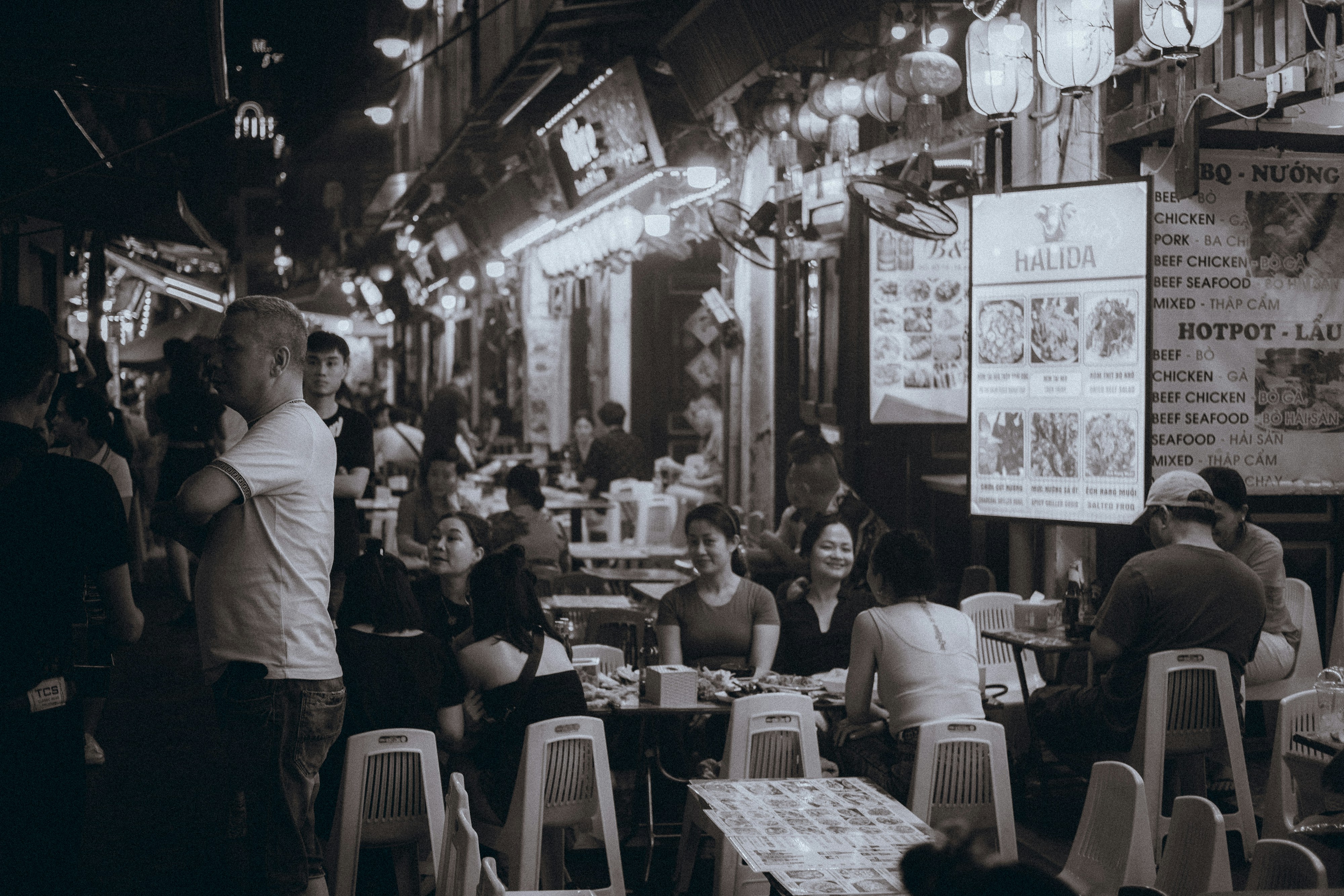 A bustling night market in Hanoi captured in black and white. The scene is filled with people sitting on small plastic chairs and tables, enjoying street food. The atmosphere is lively, with a mix of locals and tourists. Lanterns and signs hang overhead, casting a soft light over the crowded space. Food vendors are busy taking orders and serving dishes. The black and white filter adds a timeless, nostalgic feel to the vibrant market scene. The narrow street is packed with activity, highlighting the rich culture and social life of Hanoi’s night markets.