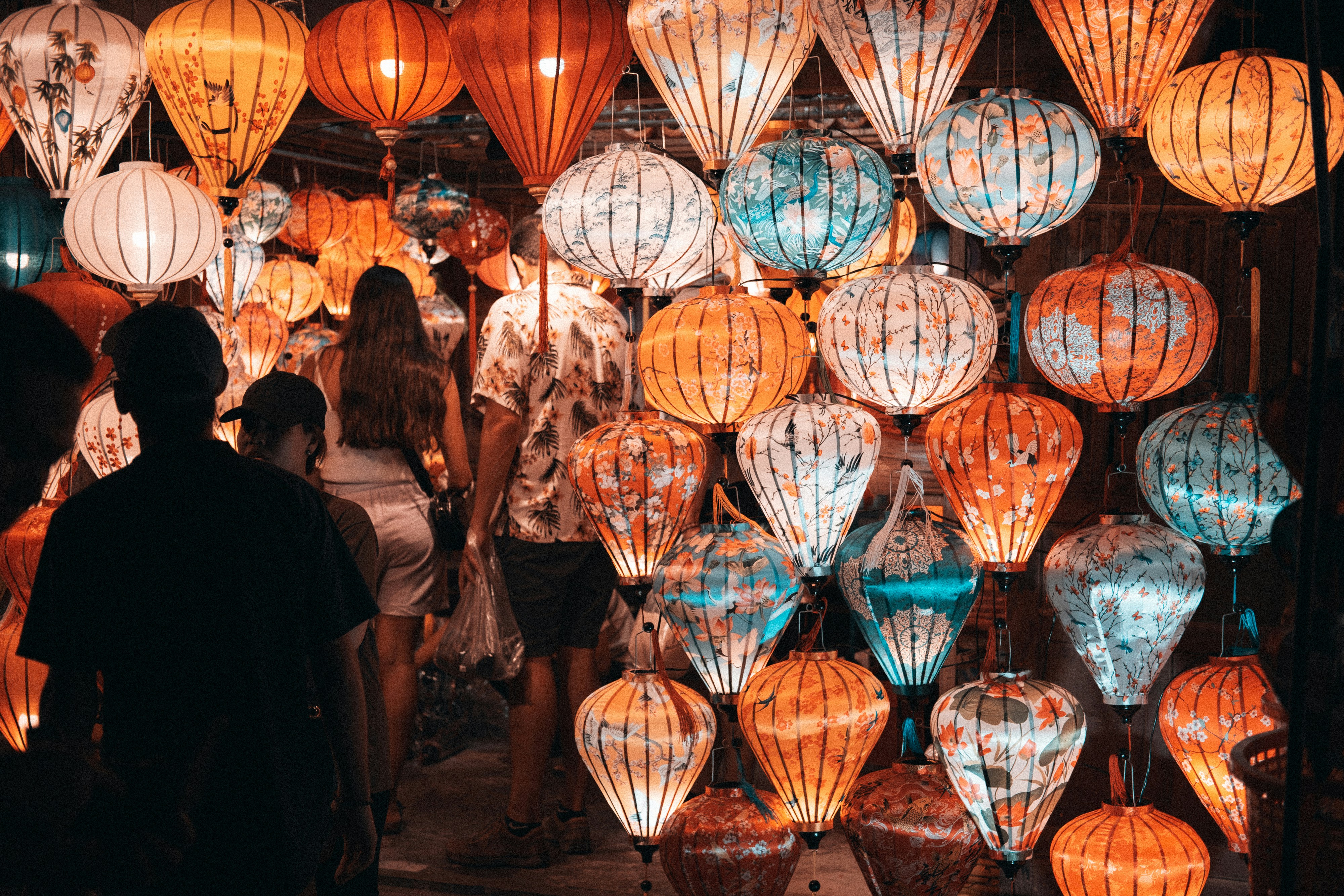 A group of people standing in front of a display of lanterns, A bustling night market in Hoi An, Vietnam, filled with people walking among a dazzling display of colorful lanterns. The lanterns, intricately designed and emitting a warm glow, create a vibrant and festive atmosphere. The scene is lively, with a mix of locals and tourists admiring the lanterns. The lanterns are primarily orange, blue, and white, hanging close together and illuminating the market with their soft light. The background is dark, making the lanterns stand out even more. The image captures the cultural richness and dynamic nightlife of Hoi An.