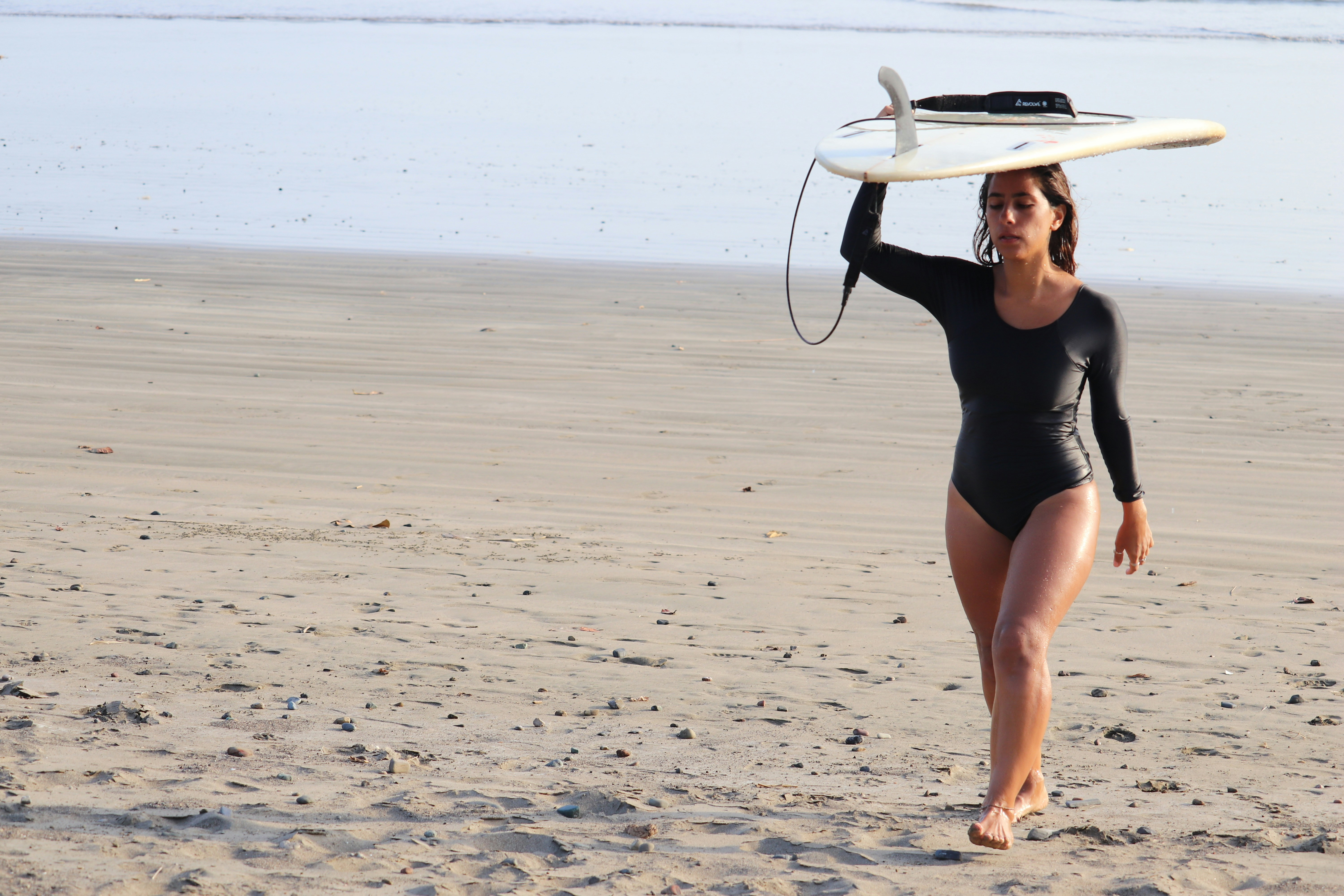 A woman in a bodysuit carrying a surfboard on a beach