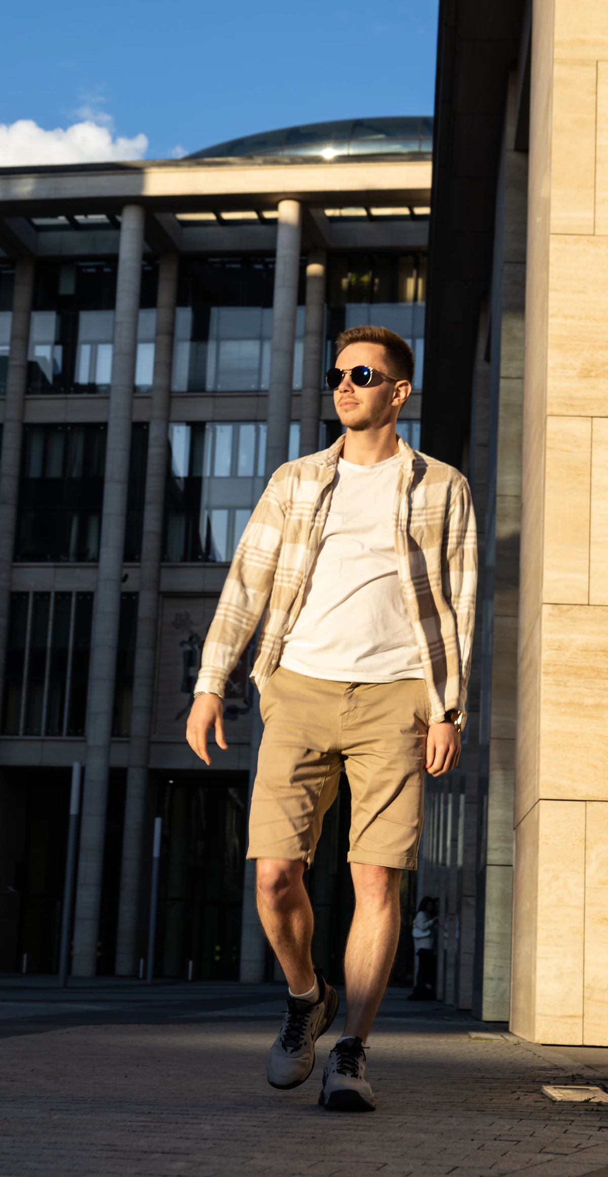 Homem de camiseta branca, camisa xadrez aberta, bermuda cáqui acima do joelho e tênis cinza caminhando em frente a um prédio