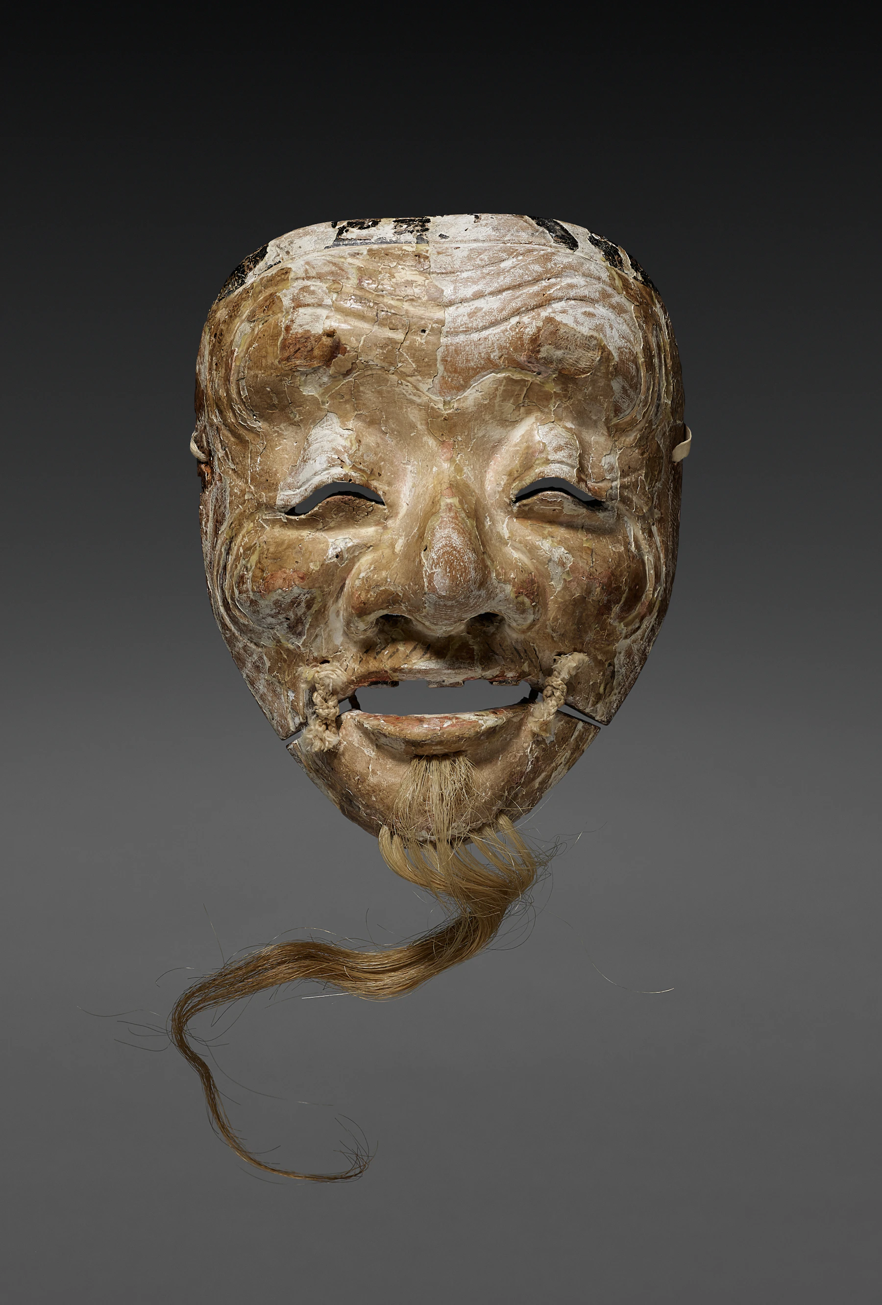 An old carved theater mask of a smiling old man with a tied-on beard, its paint worn through to the wood