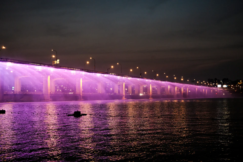 The image depicts a large, illuminated bridge spanning across a body of water at night. The bridge is adorned with purple lights that cast a warm glow on its structure and surroundings. The perspective of the photo is from the opposite side of the river, providing a view towards the bridge's end. In the background, there are buildings visible in the distance, adding depth to the scene.