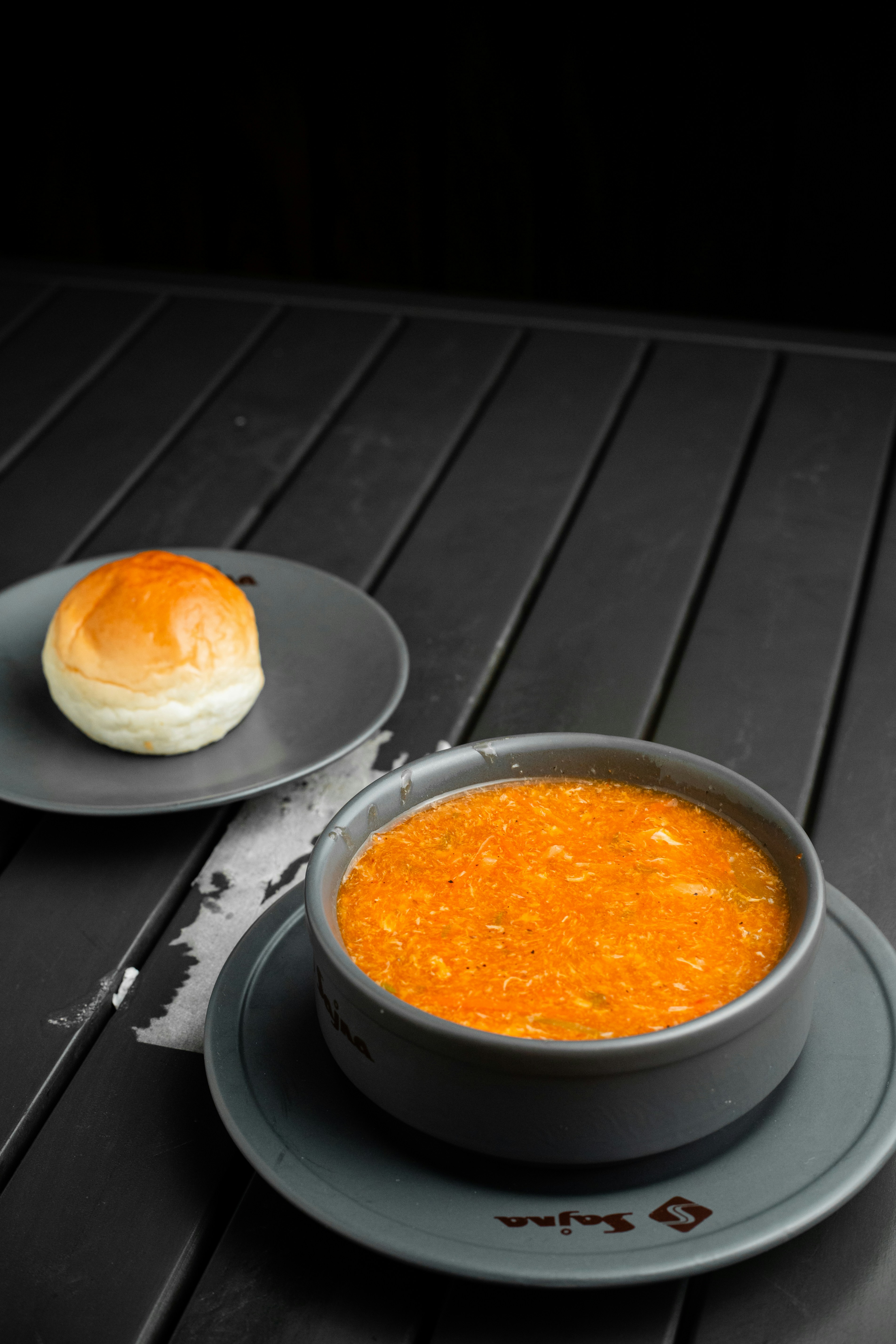 A bowl of soup next to a plate with a roll on it