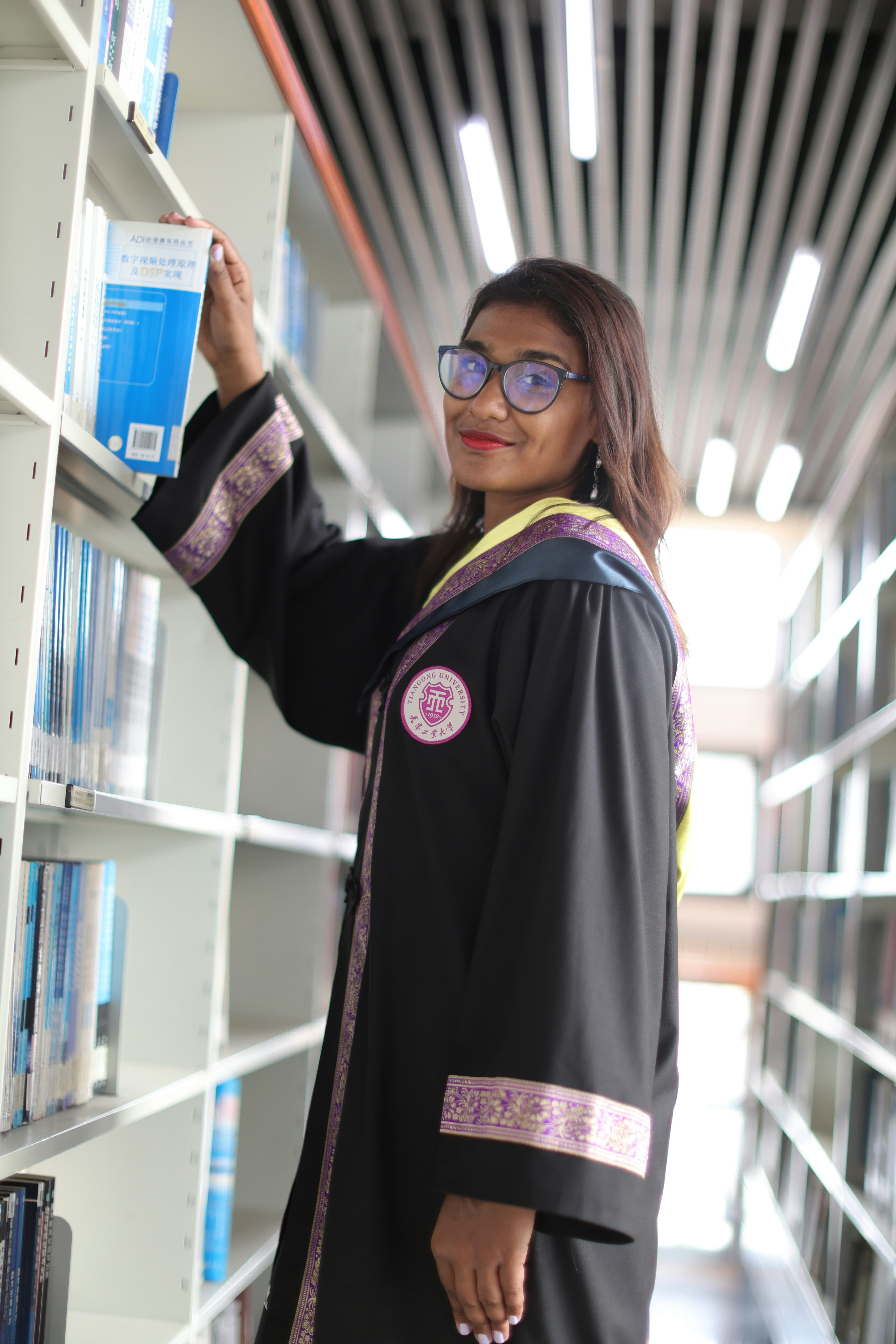 A woman in a graduation gown is looking at a book shelf
