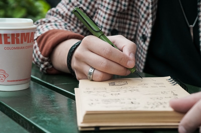 Hand holding a pen above an open notebook beside a takeaway coffee cup
