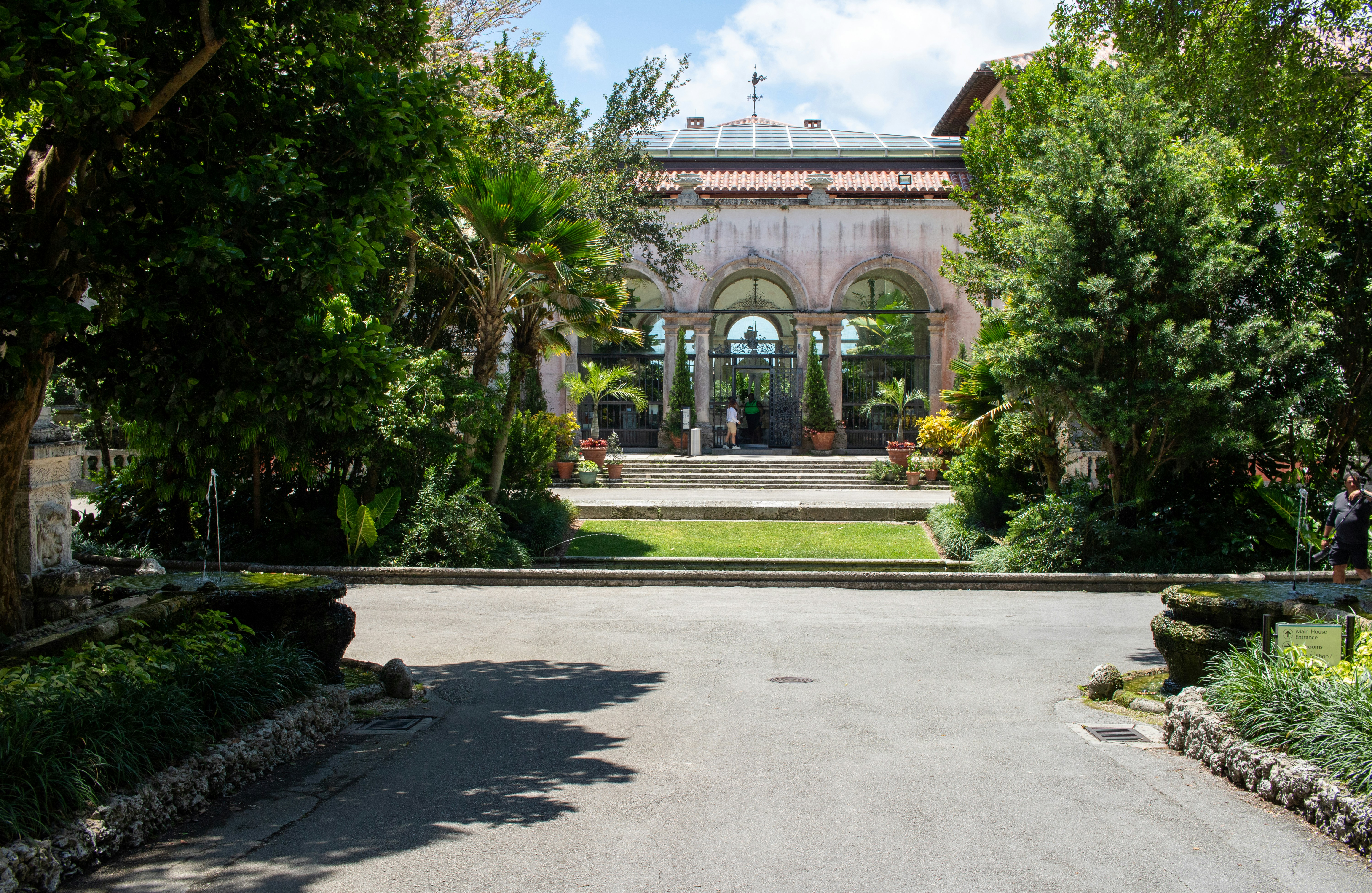 A driveway leading to a large building surrounded by trees, The Vizcaya Museum and Gardens, located in the Coconut Grove neighborhood of Miami, Florida, is an exquisite example of Mediterranean Revival architecture infused with Italian Renaissance influences. Built between 1914 and 1922 as the winter residence of industrialist James Deering, Vizcaya encompasses 34 decorated rooms filled with European antiques and art dating from the 16th to the 19th centuries. The estate originally spanned 180 acres, although today it covers approximately 50 acres, with 10 acres of formal gardens, extensive native woodlands, & a historic village outbuildings compound.