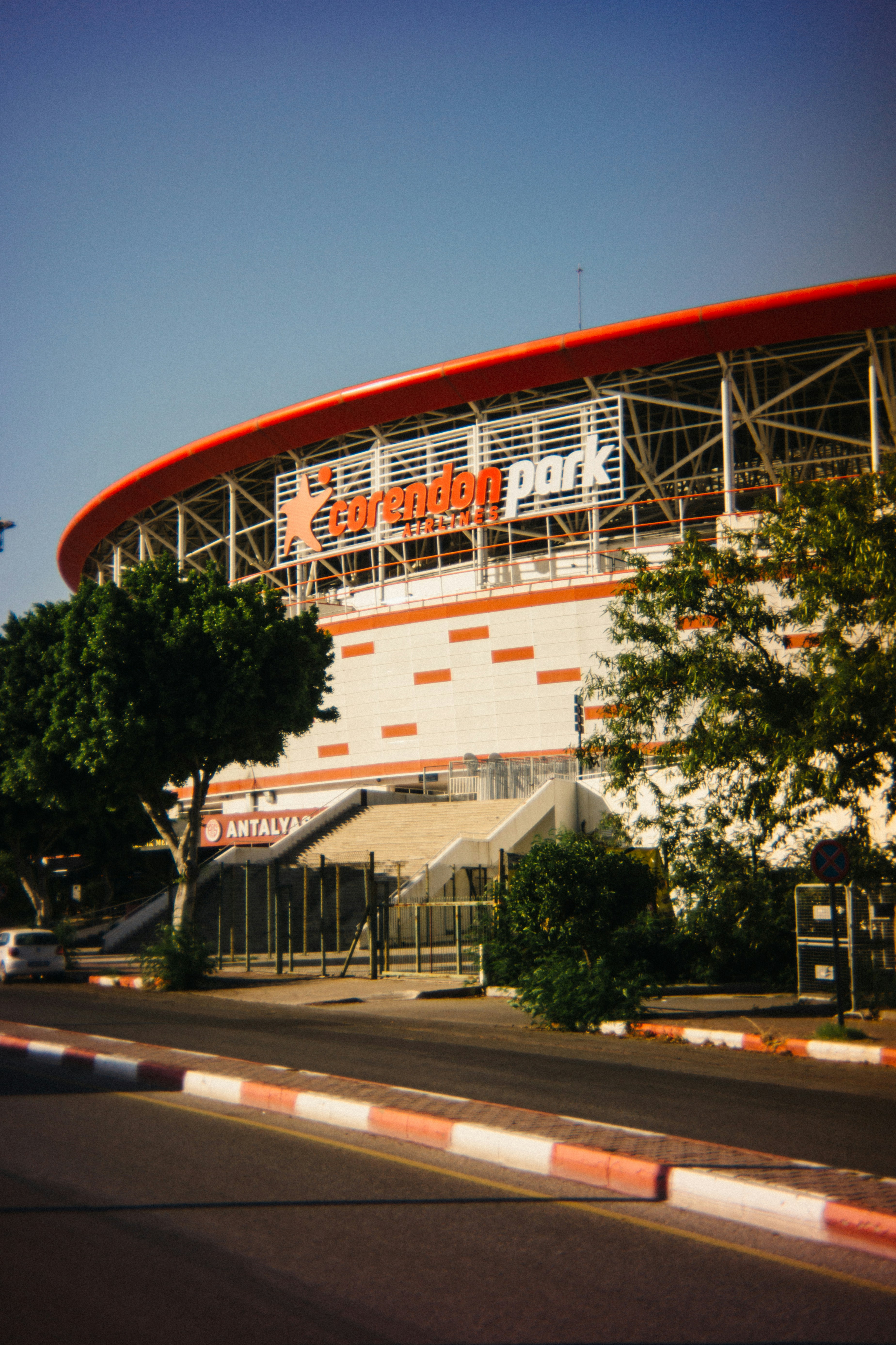 Street-level exterior view of New Antalya Stadium in Antalya, Turkey, with its red roof ring and orange-and-white facade.