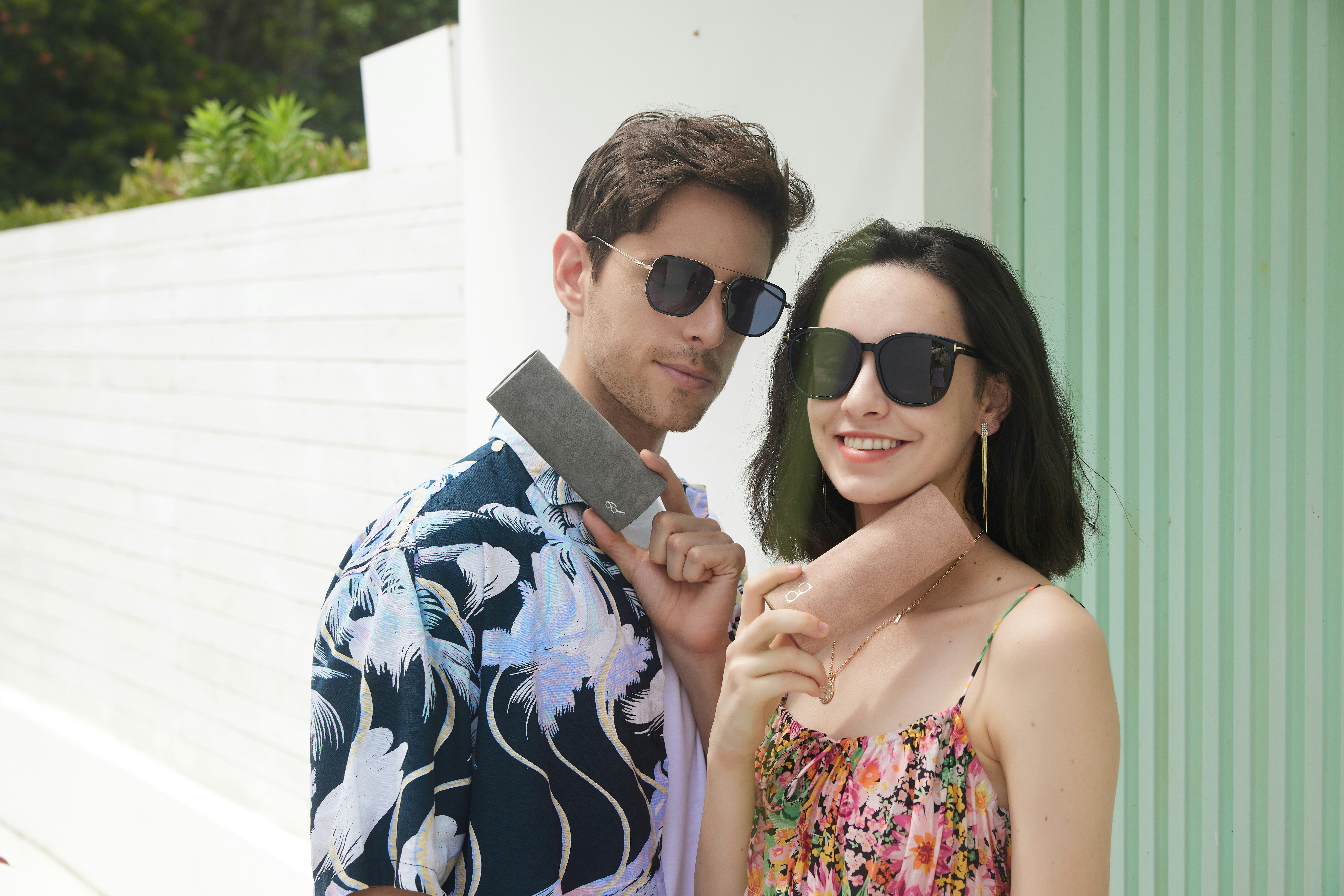 A fashionable couple enjoys a sunny day, with their stylish sunglasses taking center stage. The man wears trendy dark shades, perfectly matching his green patterned shirt. The woman, in a blue floral top, flaunts elegant cat-eye sunglasses. Their eyewear adds a touch of sophistication to their effortlessly chic look.