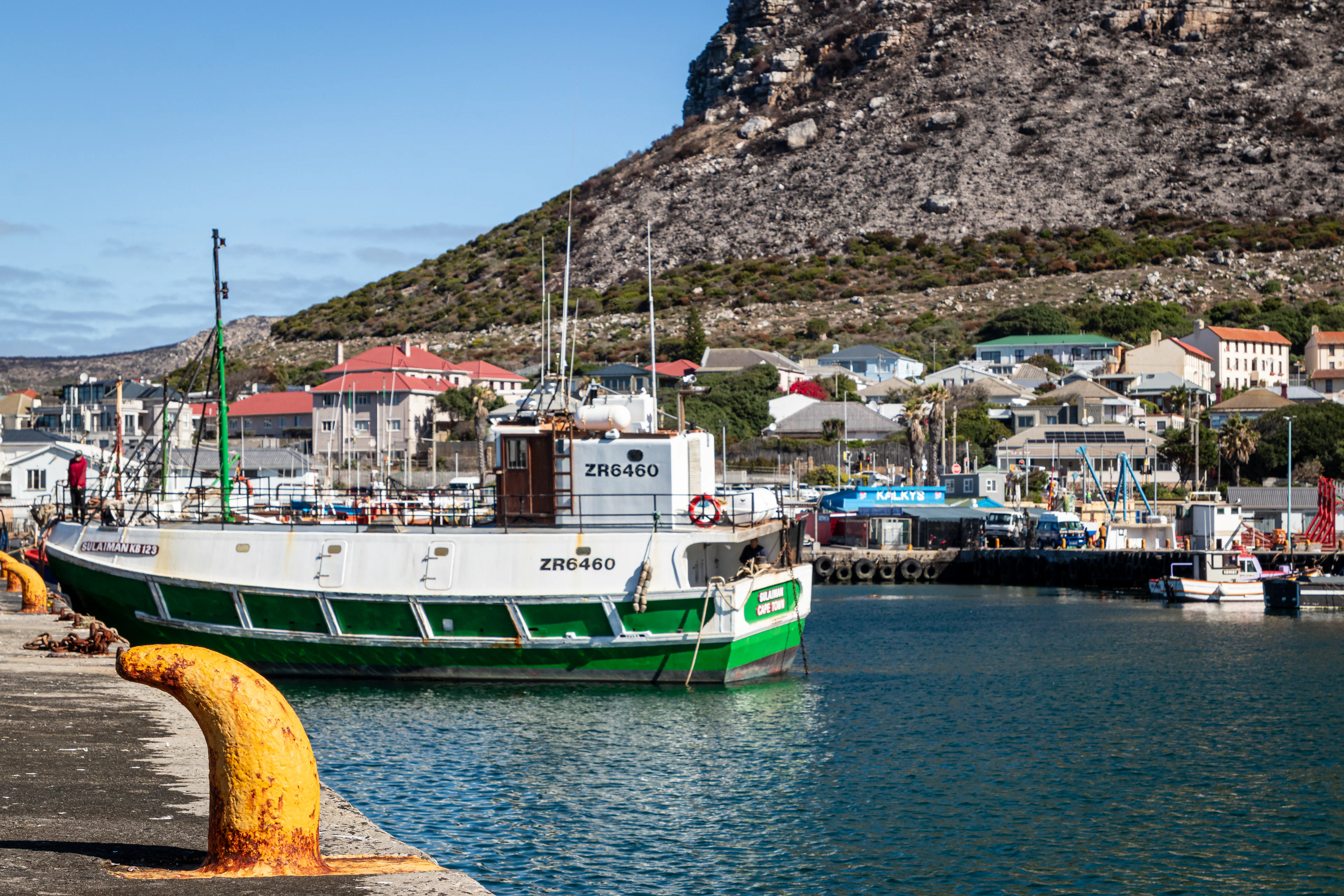 A green and white fishing trawler tied up in Kalk Bay harbour on a clear day, the painted roofs of the village and a steep brown mountain rising behind the quay