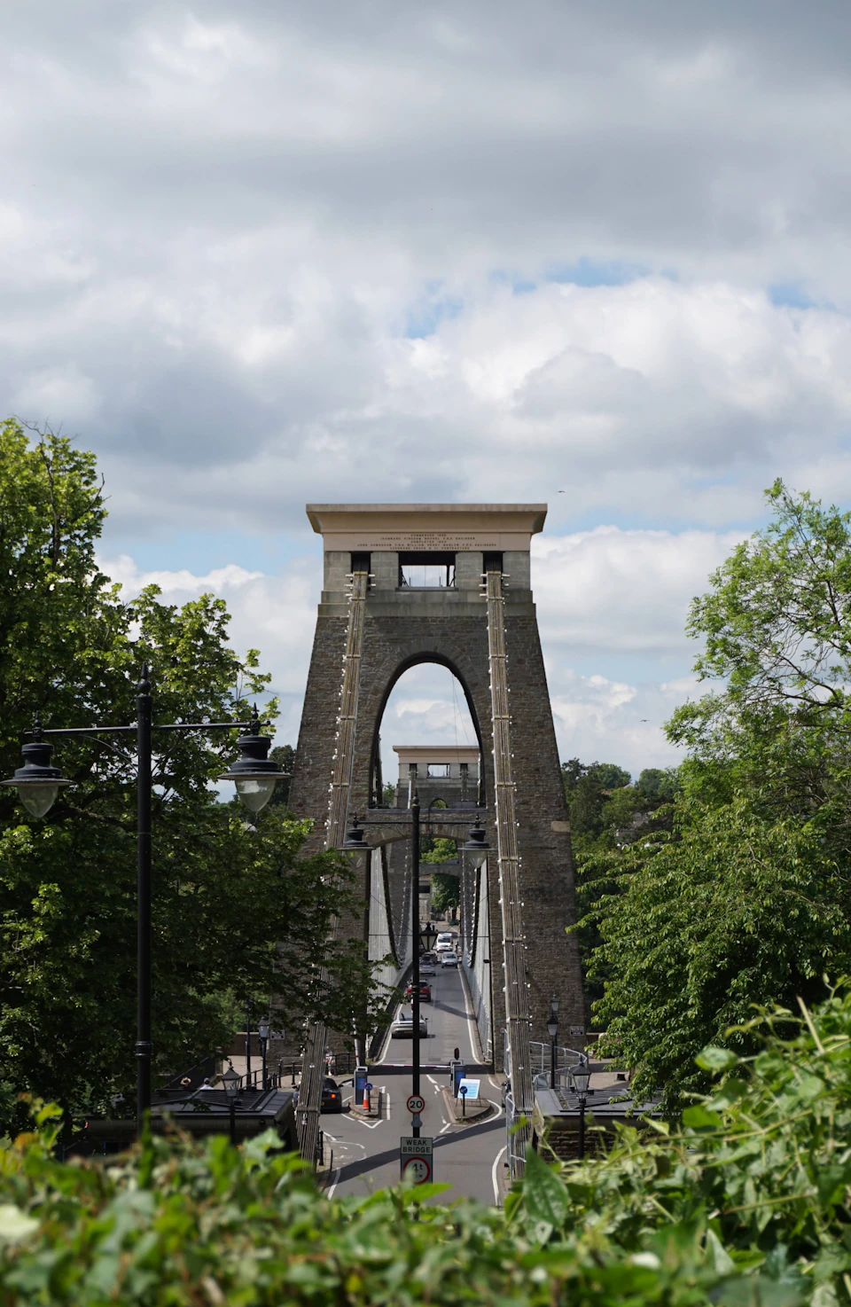 The image depicts a stone bridge with two towers, one on each side, situated over a street lined with trees and lampposts. The sky above is filled with clouds, casting a soft light over the scene. On the left side of the bridge, there are several people walking along the sidewalk, while on the right side, cars can be seen driving or parked.