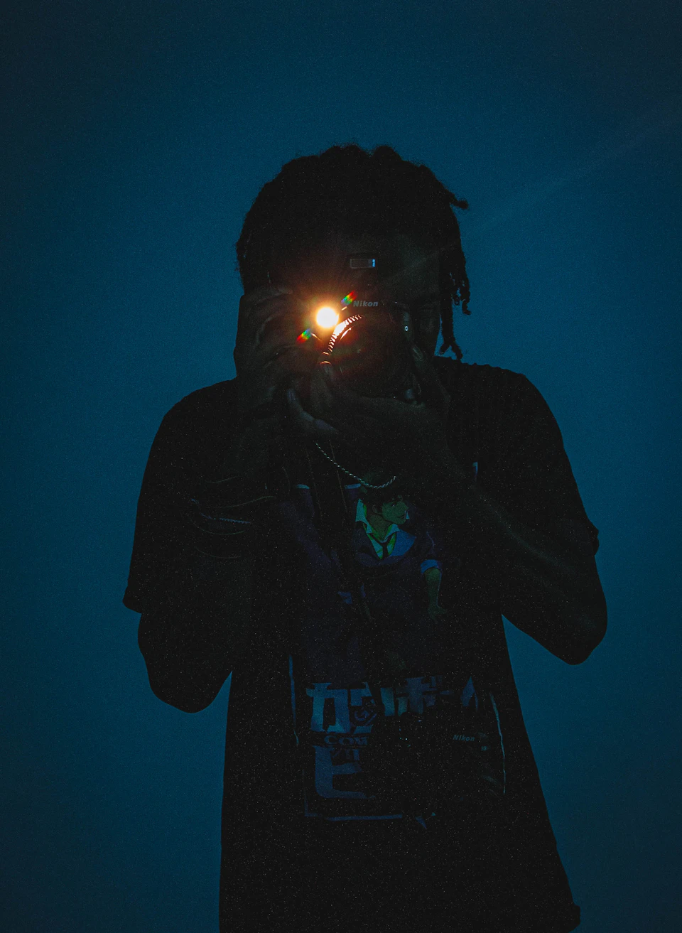 Person with dreadlocks holds up camera, capturing self-reflection in lens against dark blue background