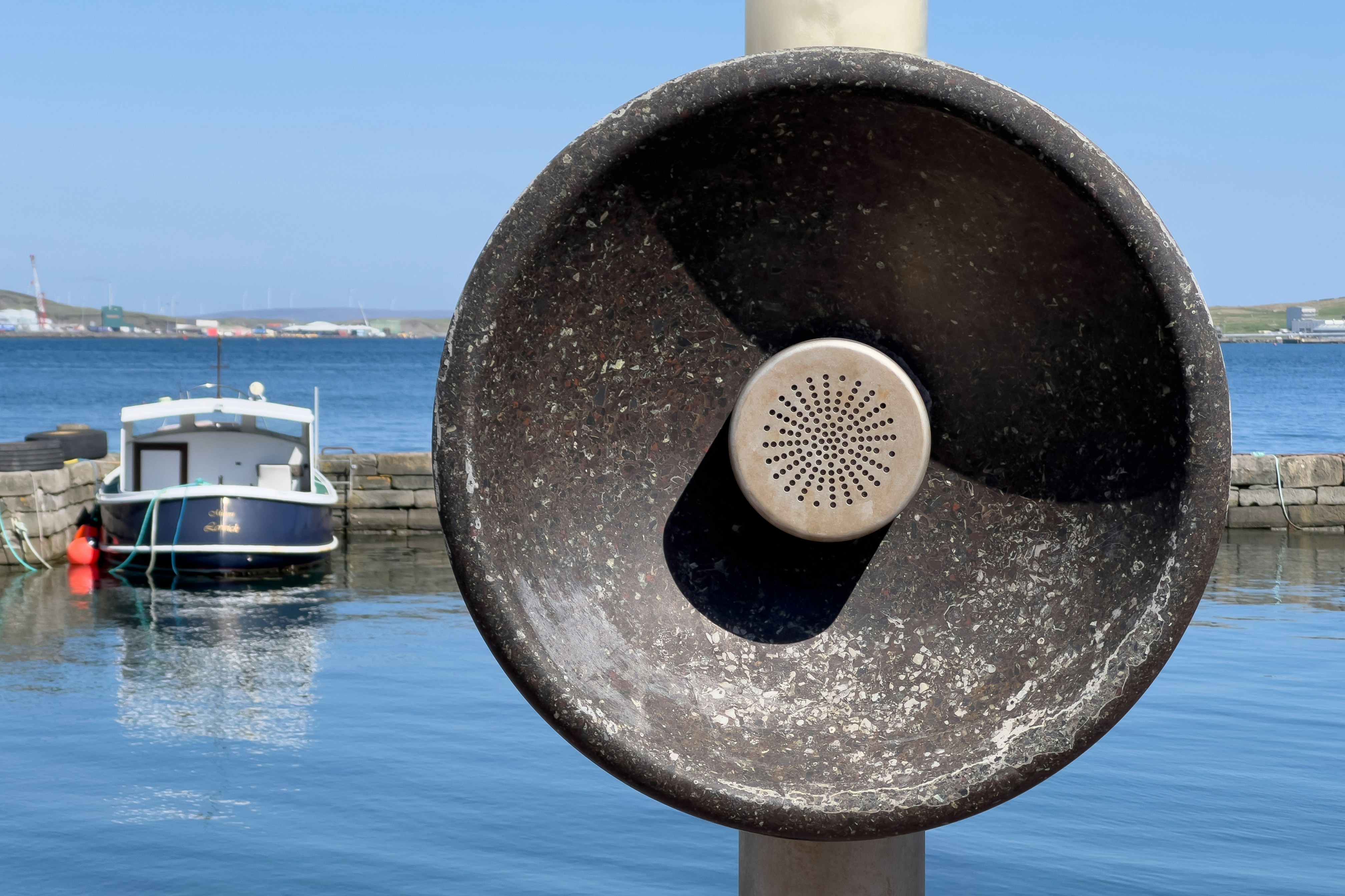 A textured, weathered sound dish stands prominently against a bright blue sky and calm harbor, with a small boat moored in the background. This unique public artwork evokes a sense of place, inviting reflection on communication and connection in a remote environment.