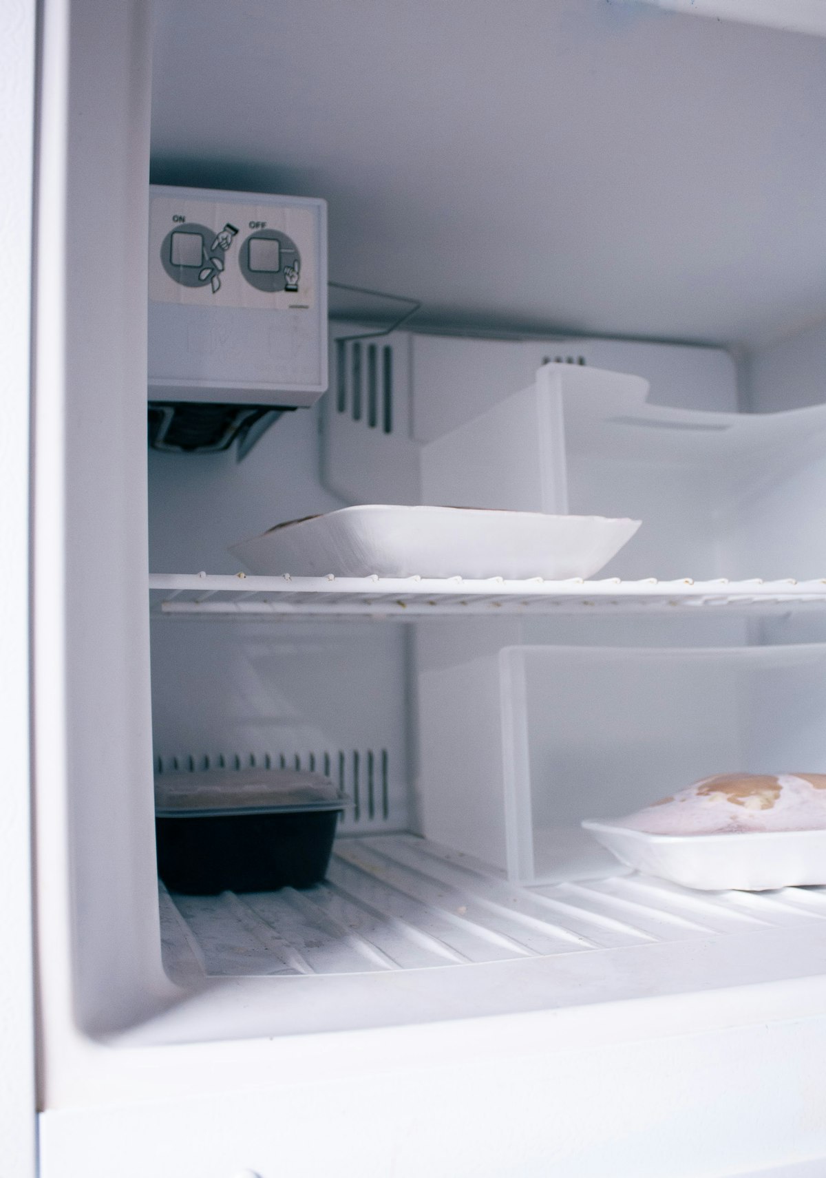 Interior de un freezer blanco abierto con estantes de rejilla, bandejas con alimentos congelados y una capa fina de escarcha sobre el piso