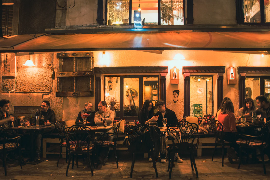 Tables full of people eating and talking outside a taverna at night, the wall behind them lit amber by wall lamps.