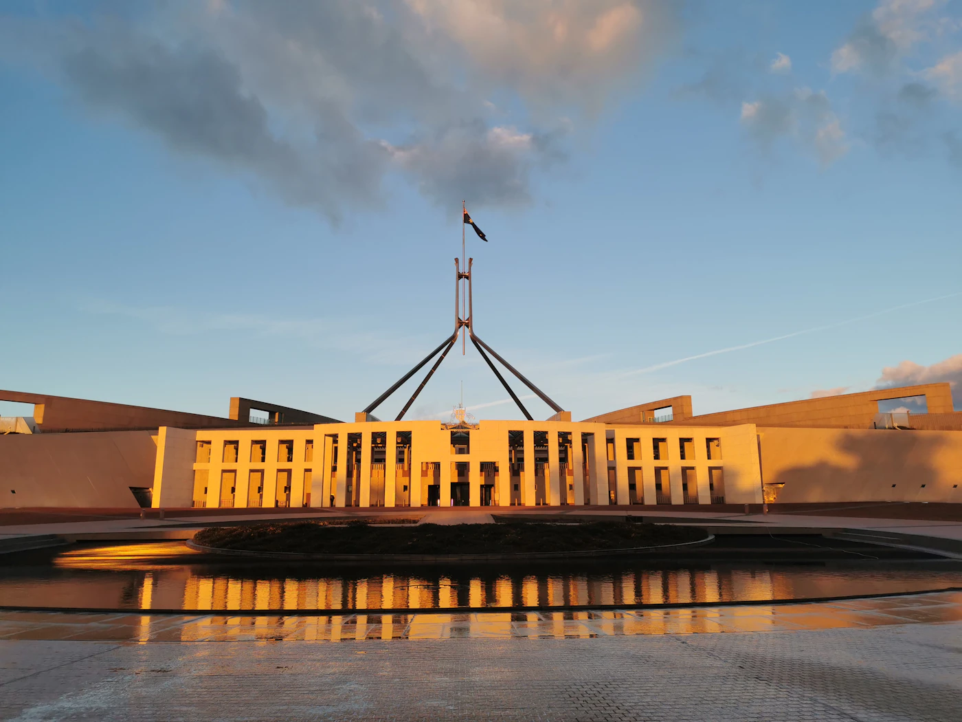 Parliament House in Canberra at sunset, its flagpole rising above the entrance, with a pool in the foreground