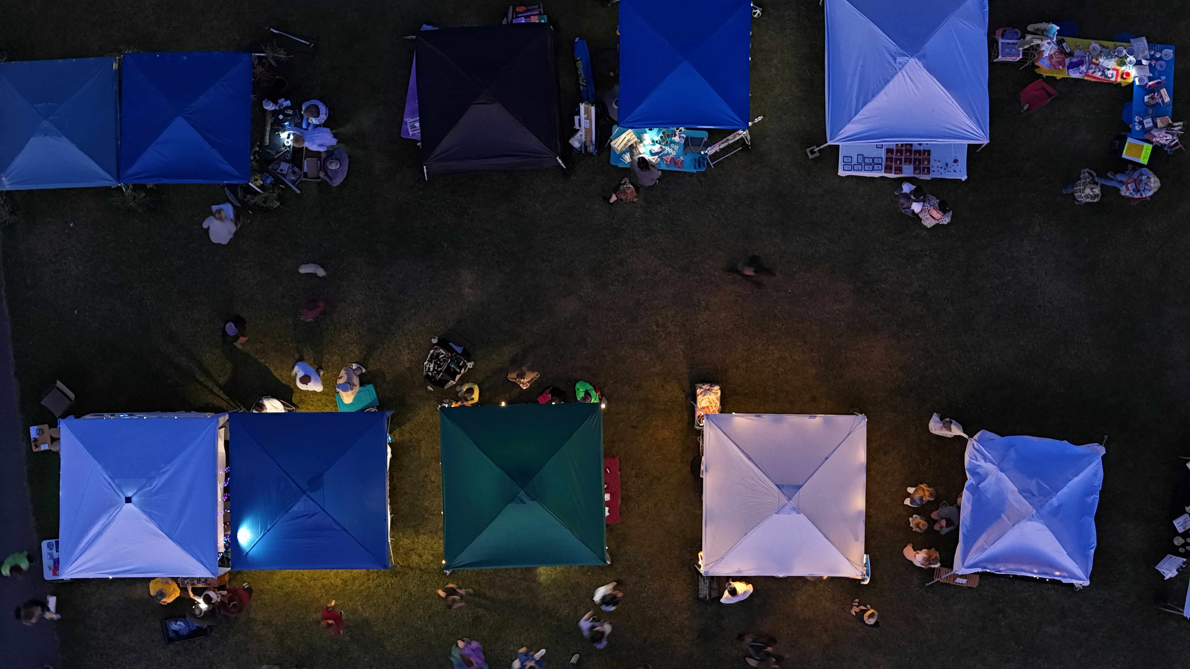 Overhead top down drone shot of an outdoor night market showing illuminated tents and people on the grass