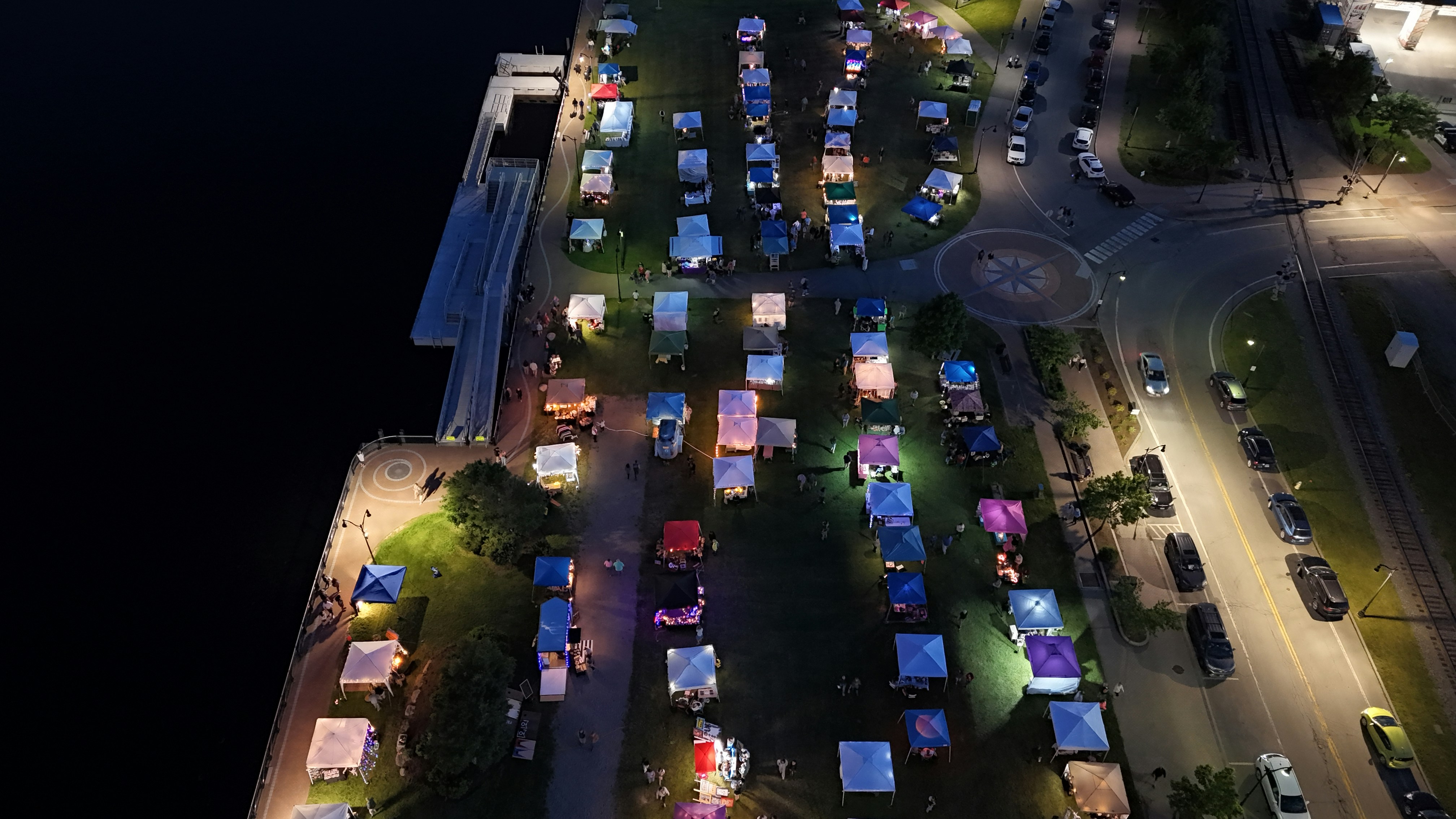 An aerial view of a parking lot at night, Drone shot of a night market showing tents and people on the grass next to a river