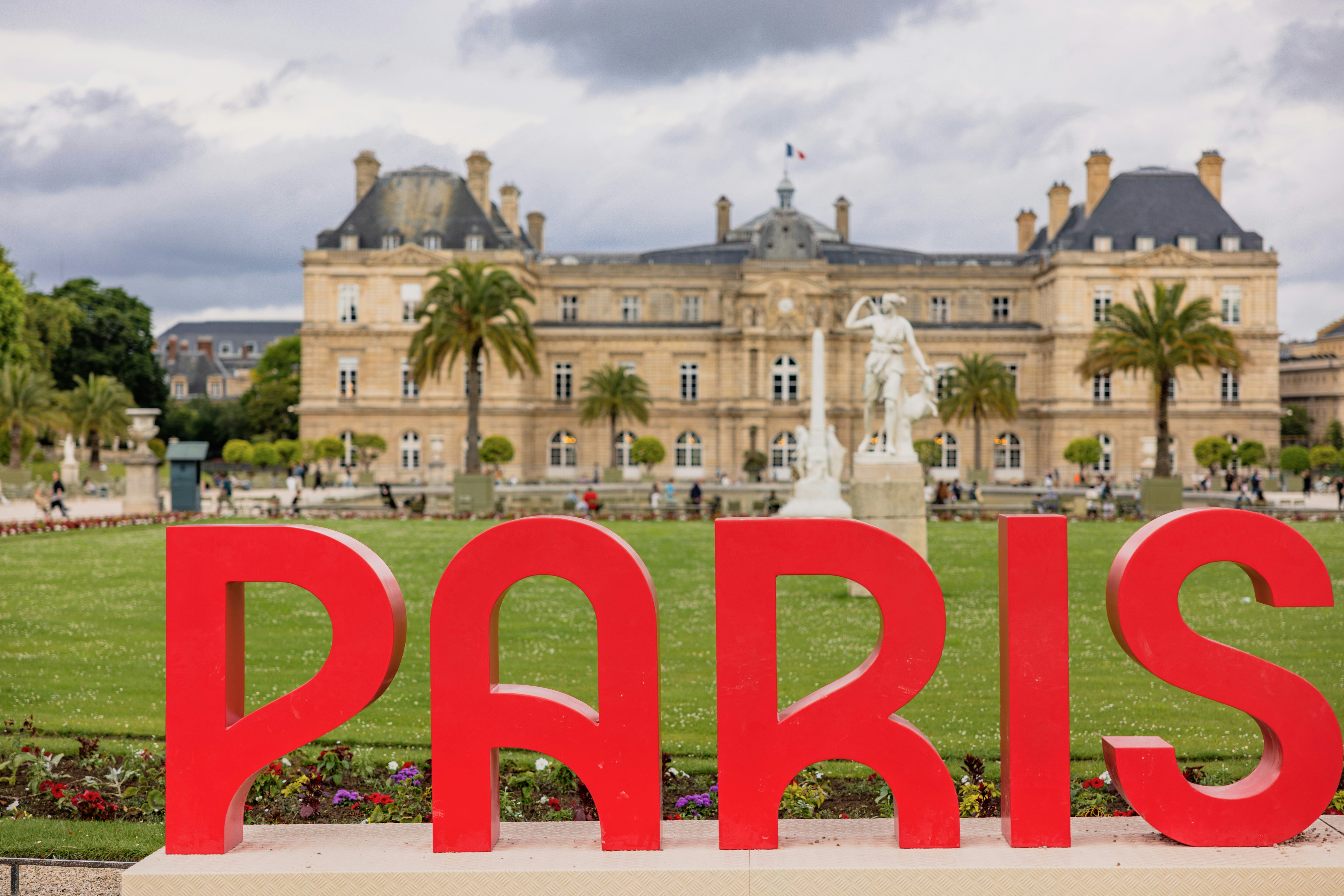 A large red sign that says paris in front of a building, A promotional installation featuring a "PARIS" logo in bold red letters, set against a backdrop of a historic palace and gardens, under a cloudy sky.