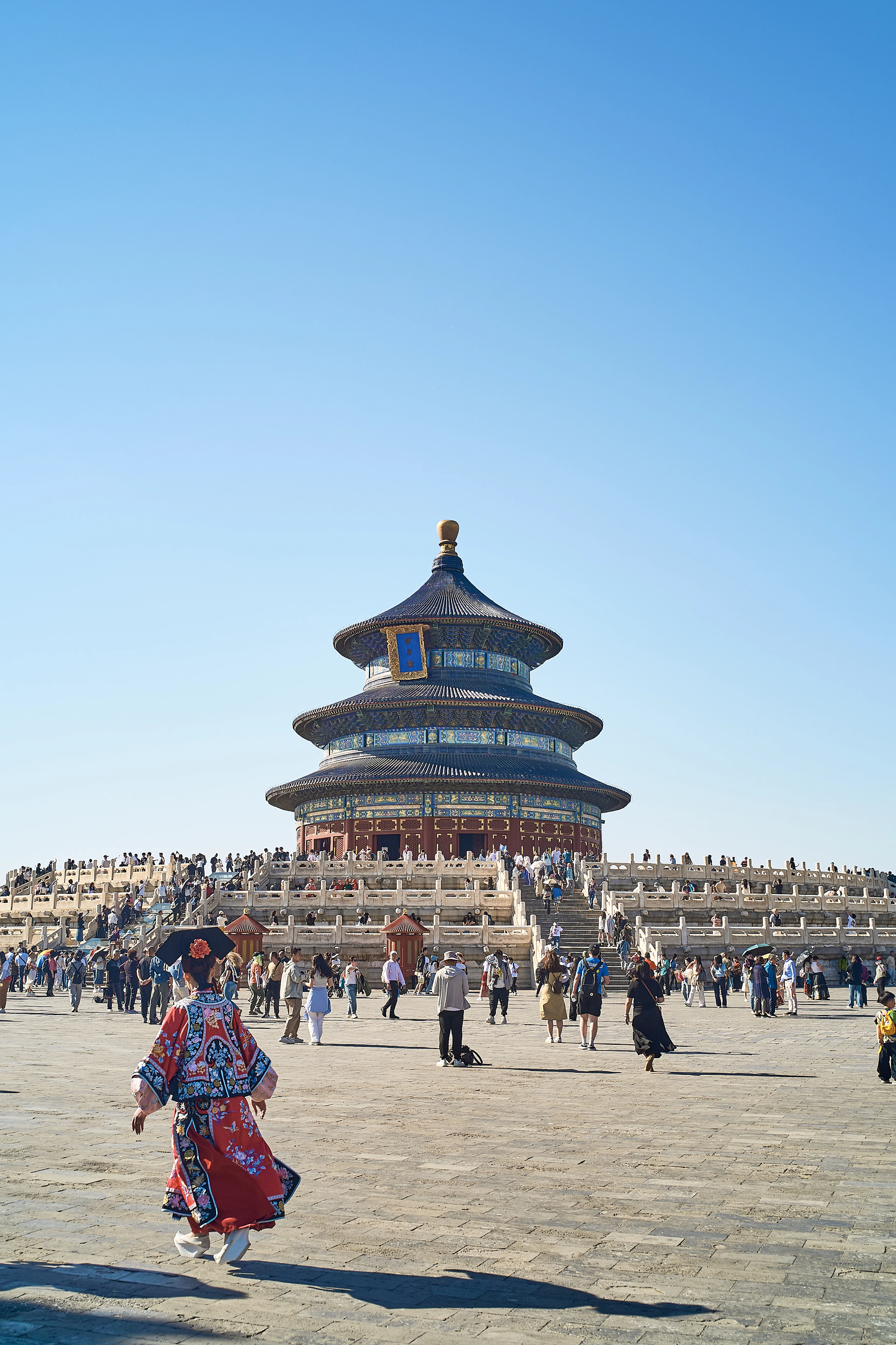 A visitor in a traditional red embroidered dress crosses the sunlit plaza before the Temple of Heaven.