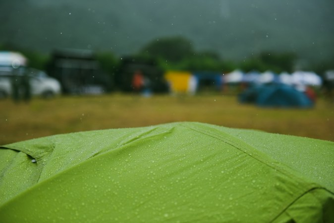 Close-up of a green tent fly beaded with raindrops, with blurred tents and parked cars in a wet field behind