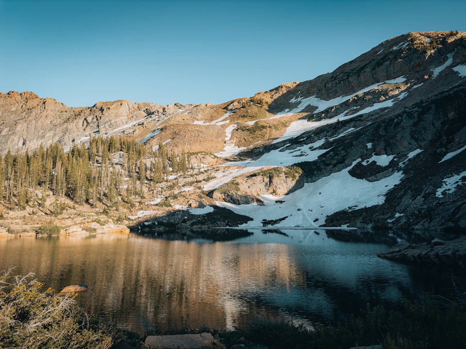 The image depicts a serene mountain lake nestled amidst a rocky landscape. The calm, dark blue water of the lake reflects the surrounding mountains and trees, creating a mirror-like effect that enhances the tranquility of the scene. On one side of the lake, there are snow-capped mountains with patches of green vegetation peeking through their icy surfaces.

The sky above is a clear blue, providing a beautiful backdrop to the natural beauty below. The image is taken from a high vantage point, offering a panoramic view of the landscape and allowing for an appreciation of the grandeur of nature's creations.