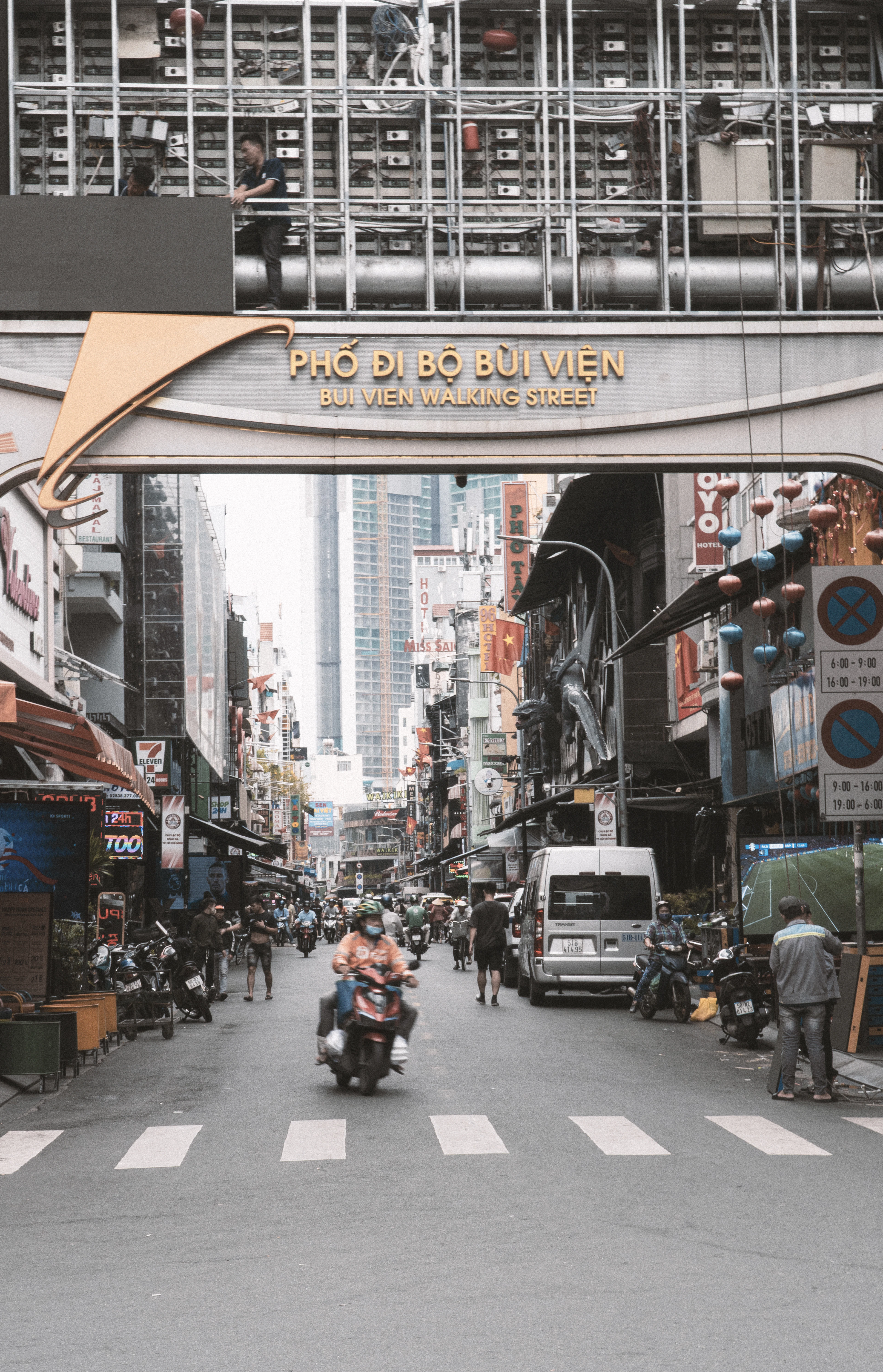 A moped rider crosses Bui Vien Walking Street in Ho Chi Minh City beneath its arched bilingual sign and red lanterns at dusk.
