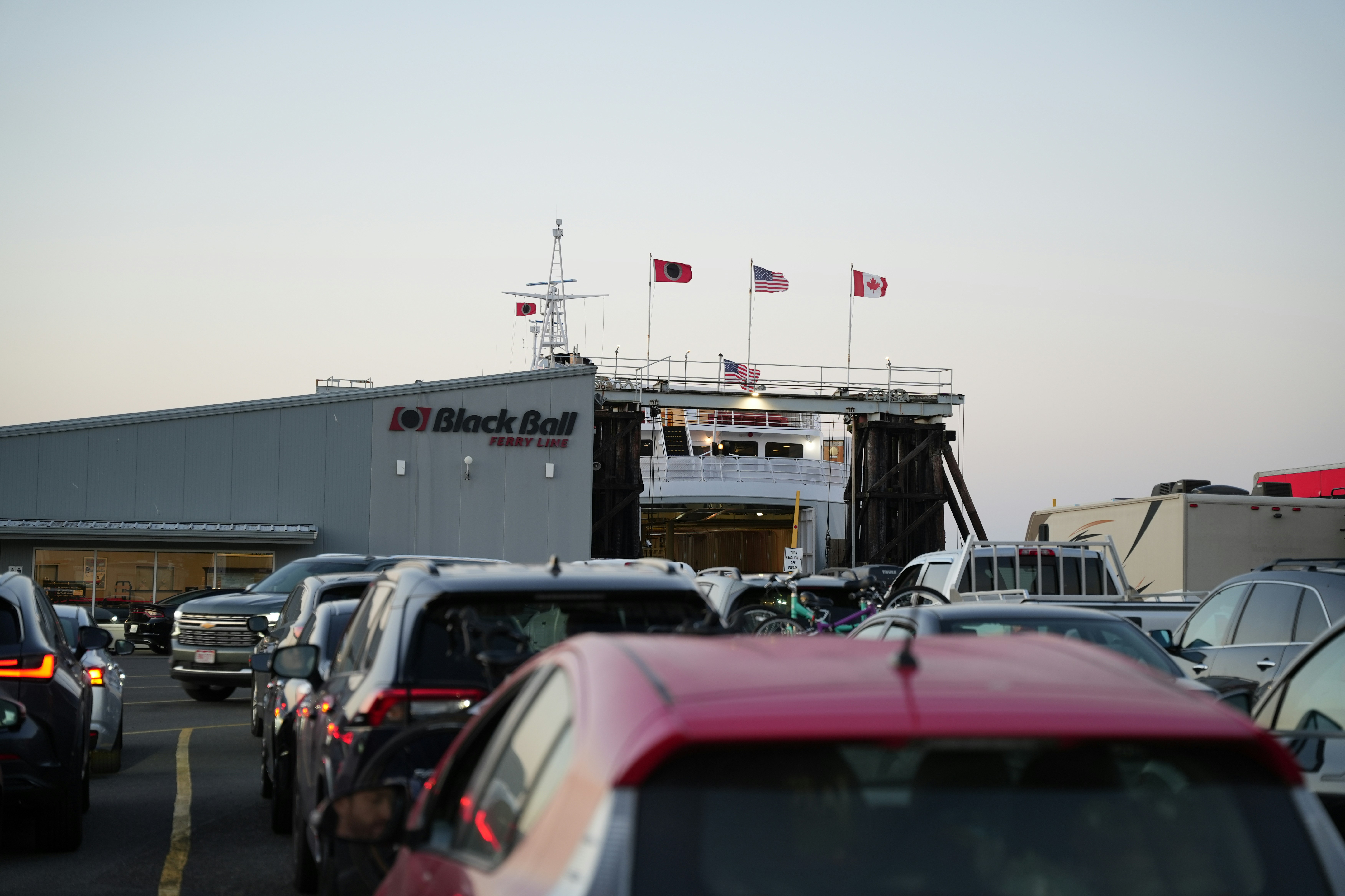 A busy highway filled with lots of traffic, Vehicles waiting to board the ferry at Black Ball terminal during dusk, flags waving in the soft evening light.