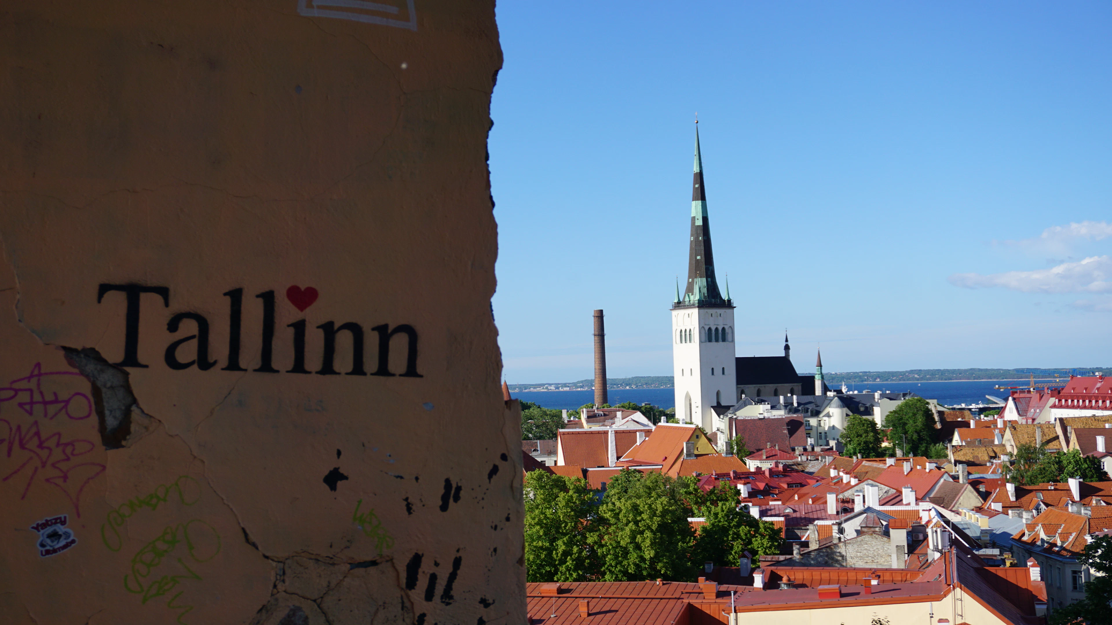 Tallinn's red roofs and church spire from a viewpoint, the city's name painted on the wall in front