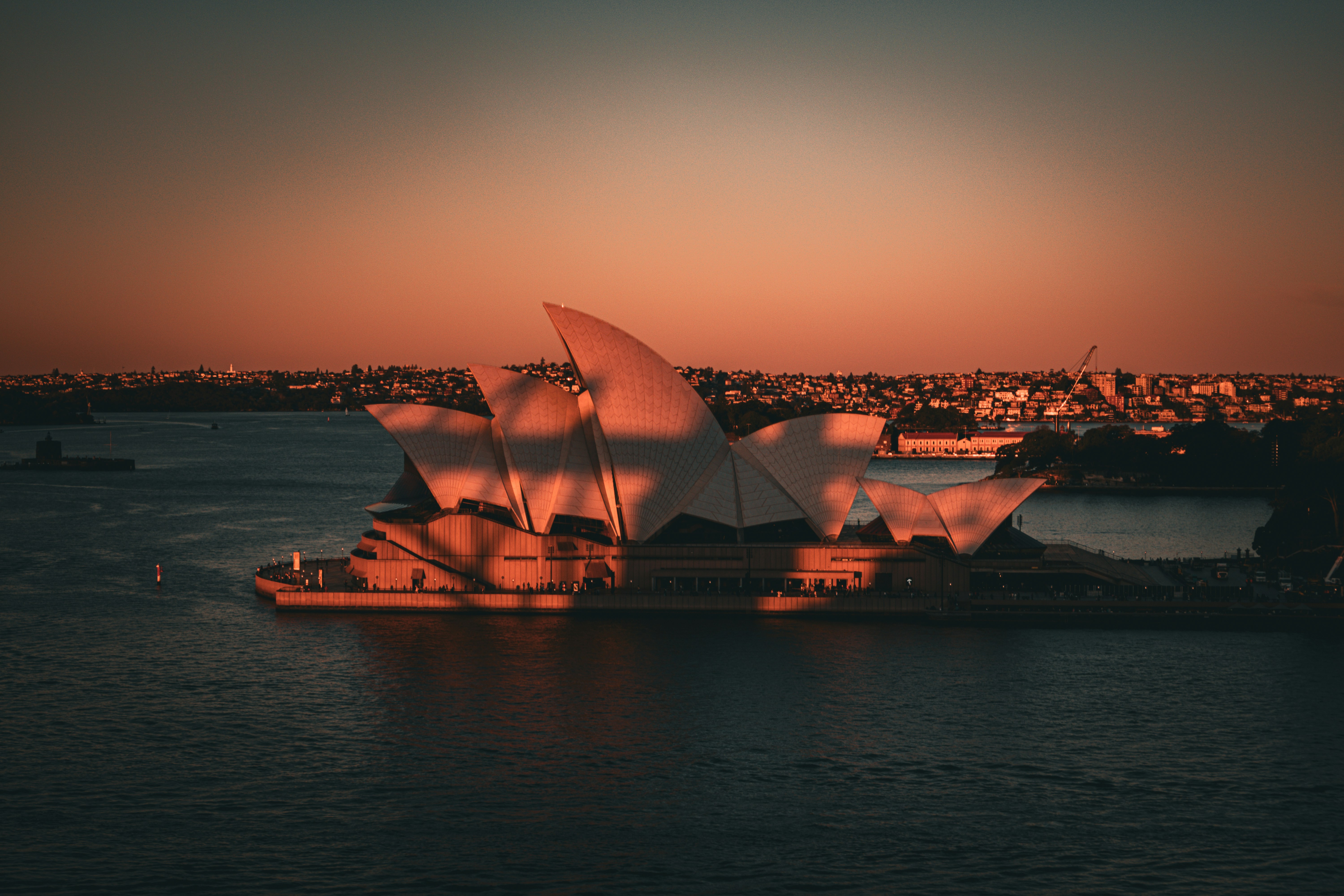A large boat in the water near a city, The Sydney Opera House is illuminated by the soft, warm light of the setting sun, casting a golden glow on its sail-like structure. The surrounding water and distant cityscape are bathed in the same rich hues, creating a serene and breathtaking view at dusk.