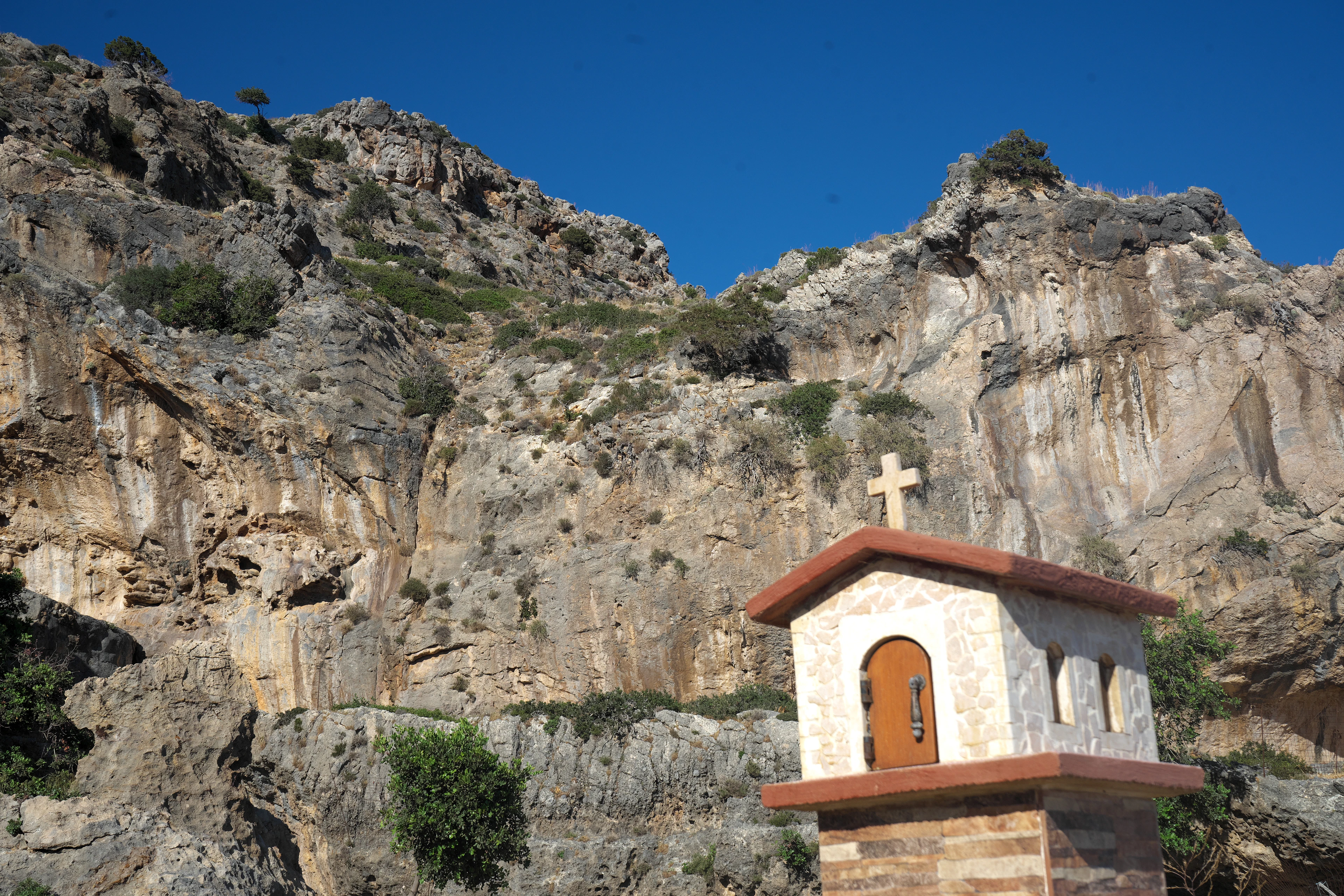 A small whitewashed roadside chapel with a cross stands against a sheer limestone cliff in Crete.