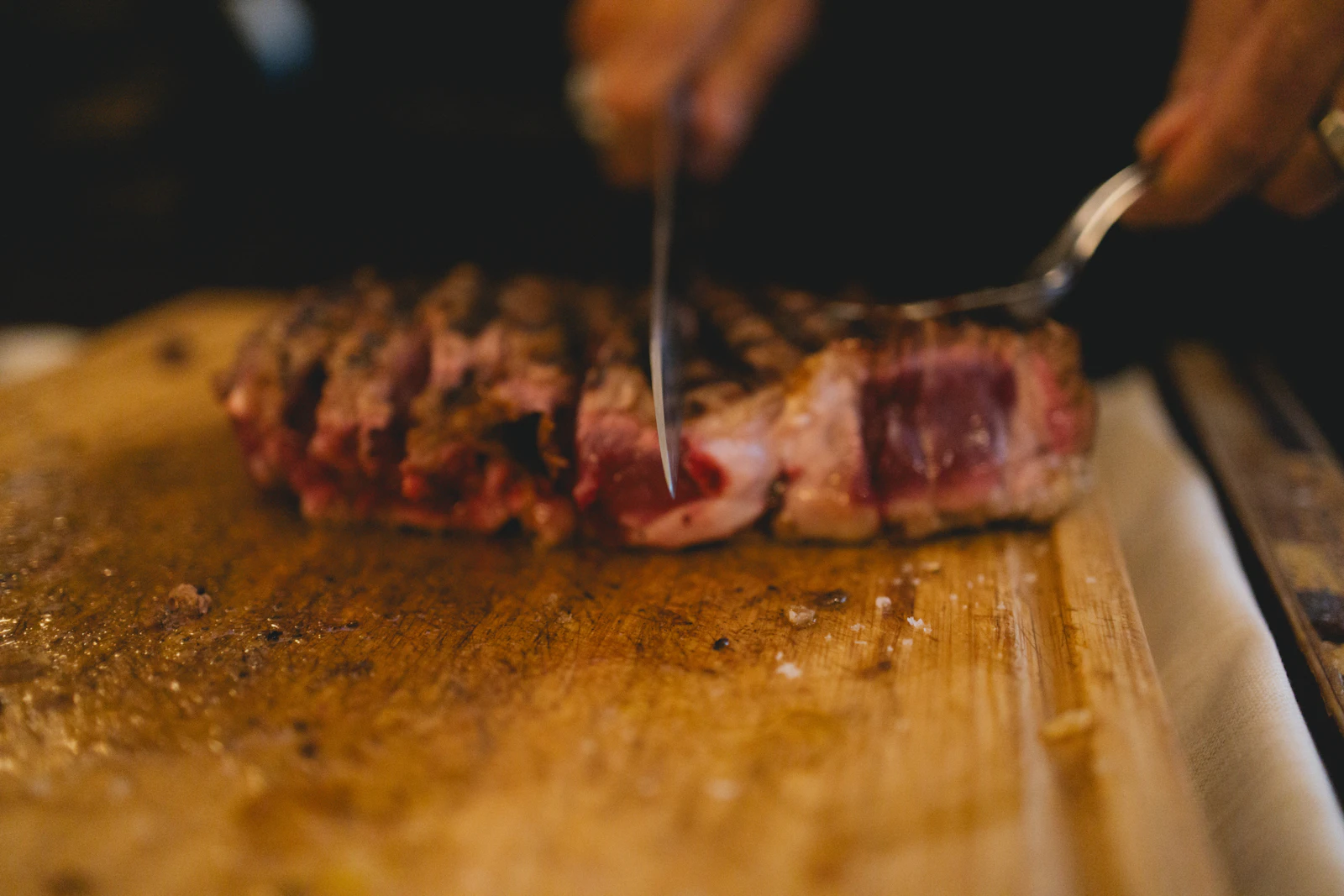 Hands slicing a seared steak on a wooden board with a carving knife and fork