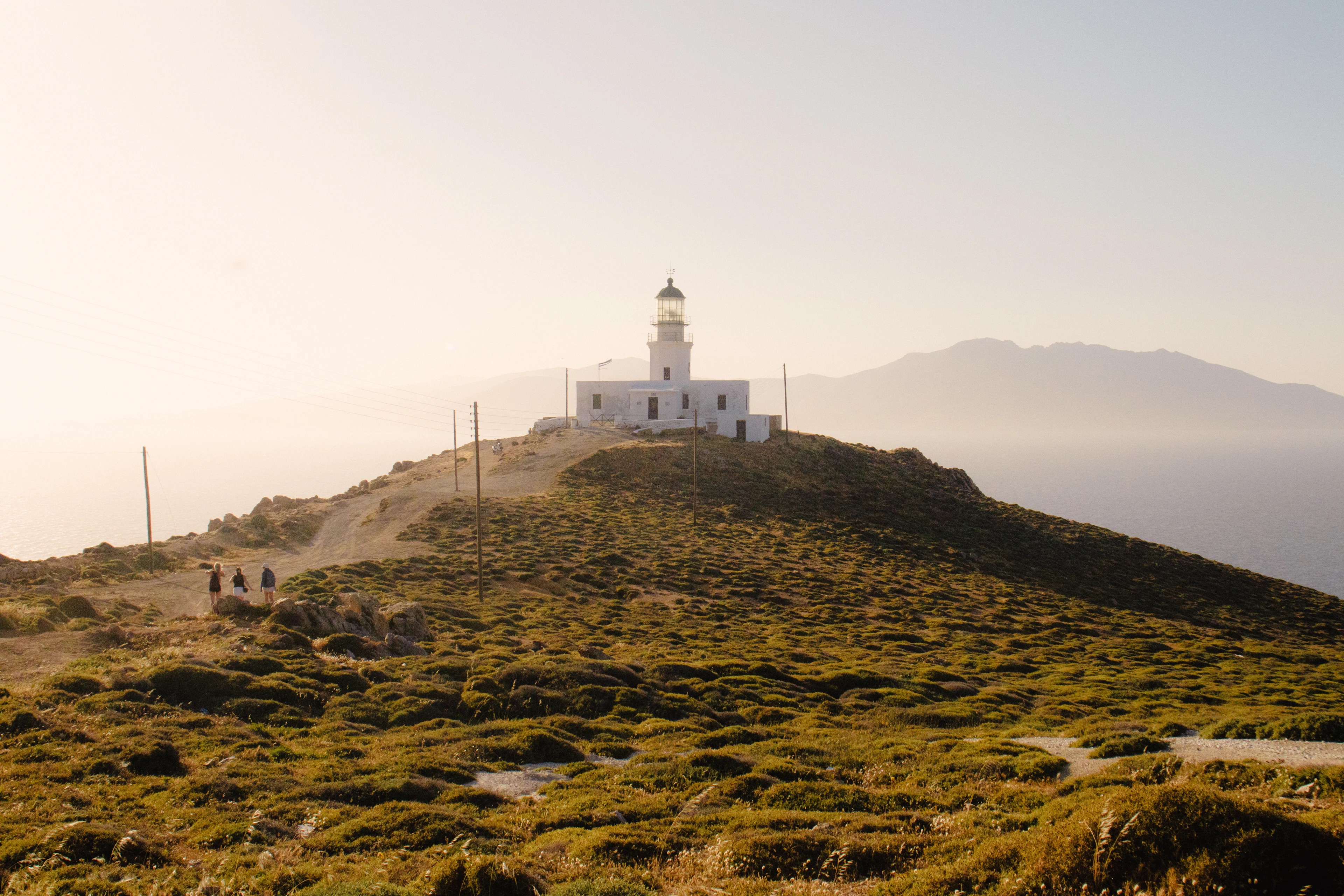 The white Armenistis lighthouse standing on a green, moss-soft headland at the northern tip of Mykonos, walkers on the path below it and a hazy blue island ridge across the water in low golden light.