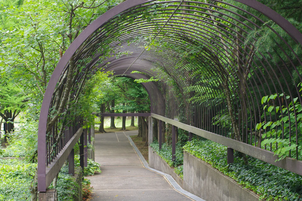 The image depicts a serene park scene with a curved metal archway constructed from green metal, supported by two sturdy columns on either side and adorned with lush trees and bushes. The archway is situated within a concrete walkway that extends into the distance, leading to a small pond in the background. The sky above is overcast, casting a soft light over the scene.